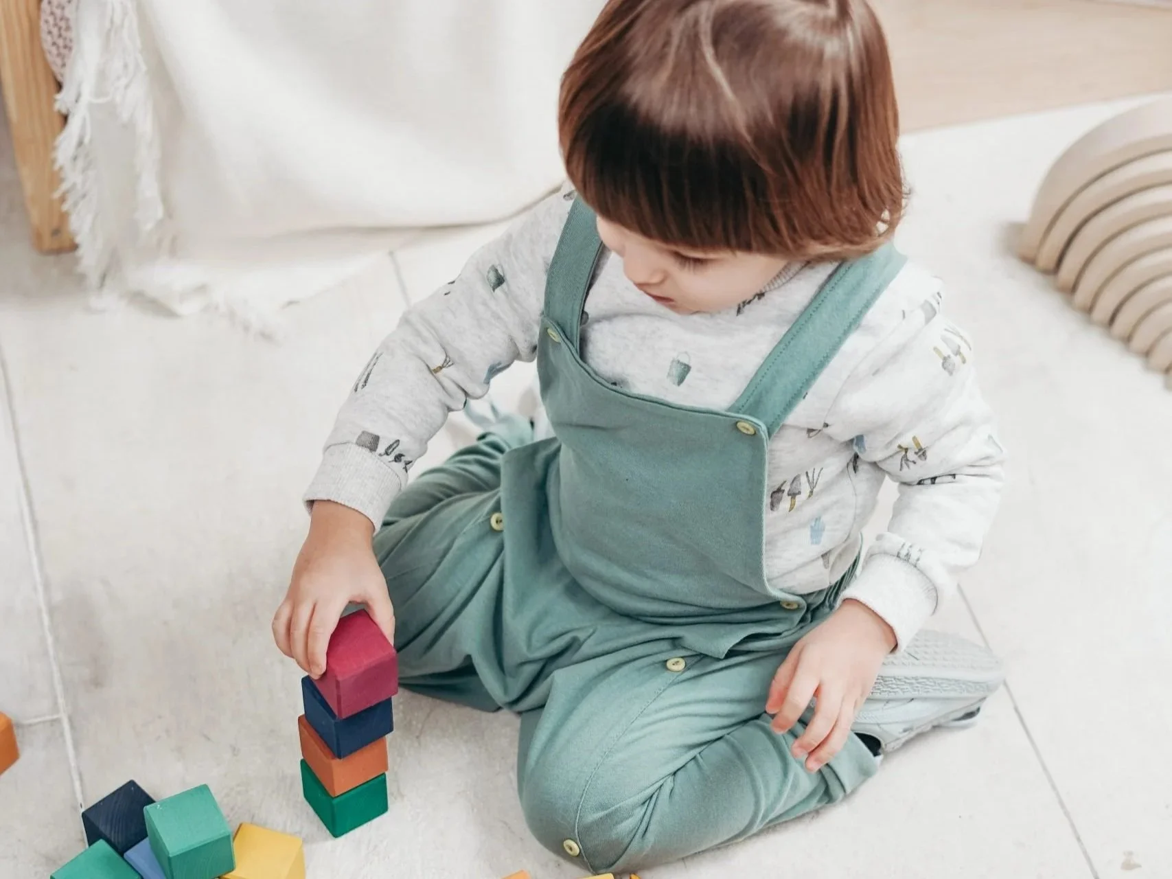 A child in green overalls sits with their legs in a 'w' position while stacking blocks