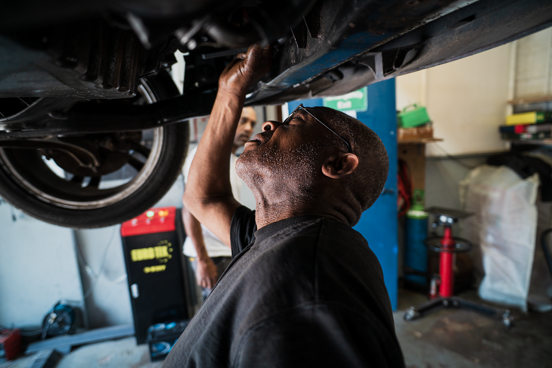 Man working underneath a lifted car in a garage, inspecting or repairing the vehicle.