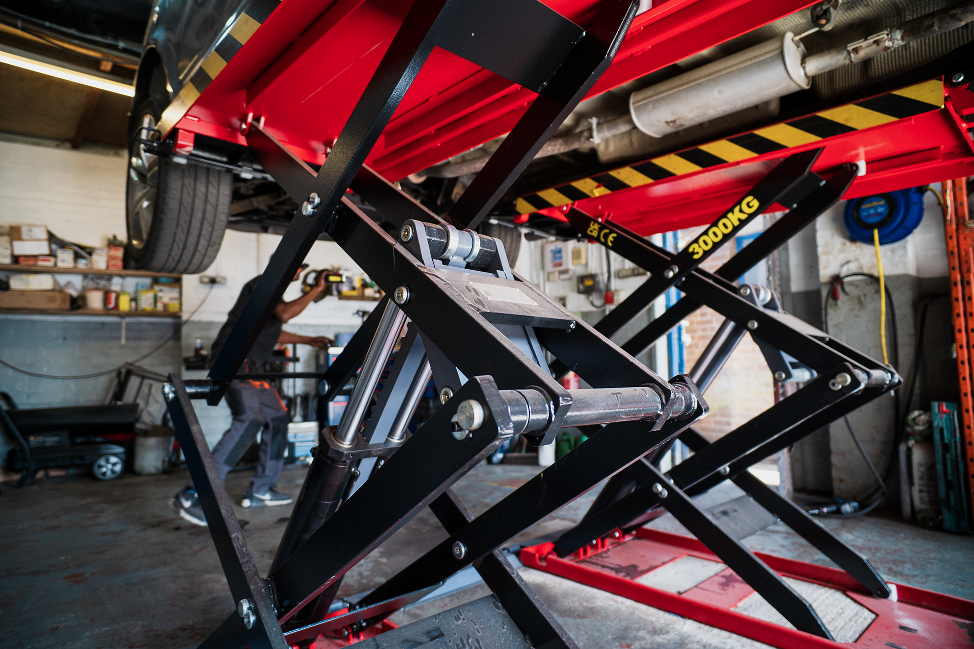 Underneath view of a race car on a hydraulic lift in a garage, showing the underside details, with a mechanic working in the background.
