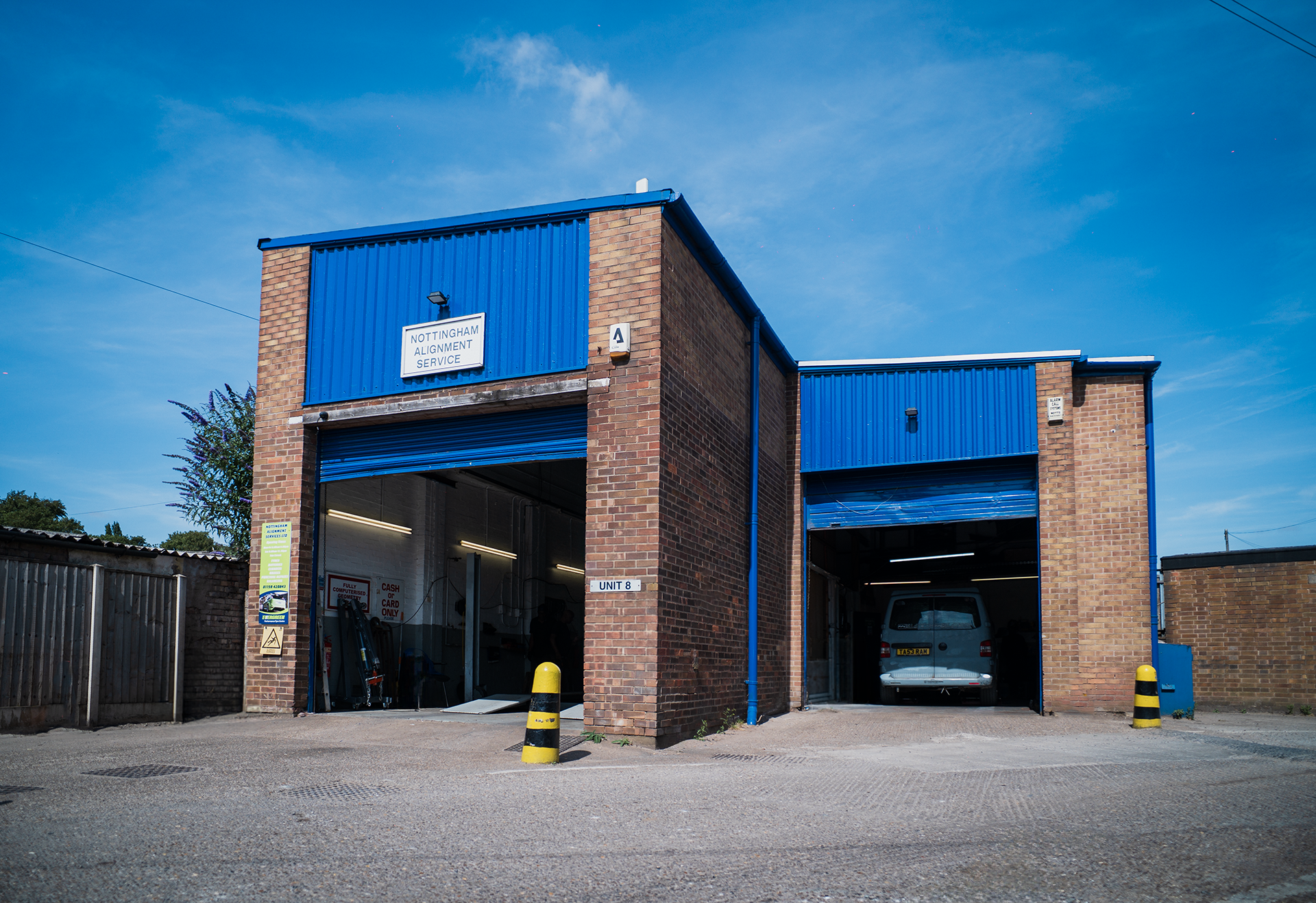 A brick and blue metal building with two large garage doors, one open revealing a vehicle inside, and a sign reading Nottingham Alignment Service on the front. There are yellow and black safety bollards in front of the entrance.