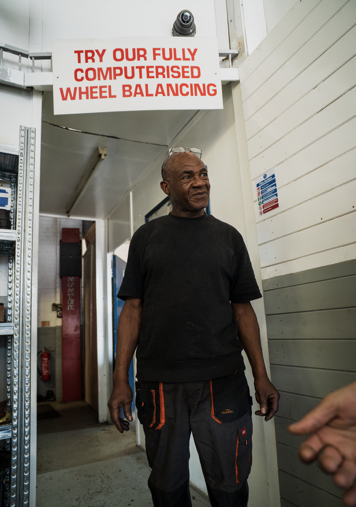 A man standing indoors near a gray wall and a doorway, wearing black and orange work pants and a black t-shirt, with a sign above him that says 'Try our fully computerised wheel balancing.'