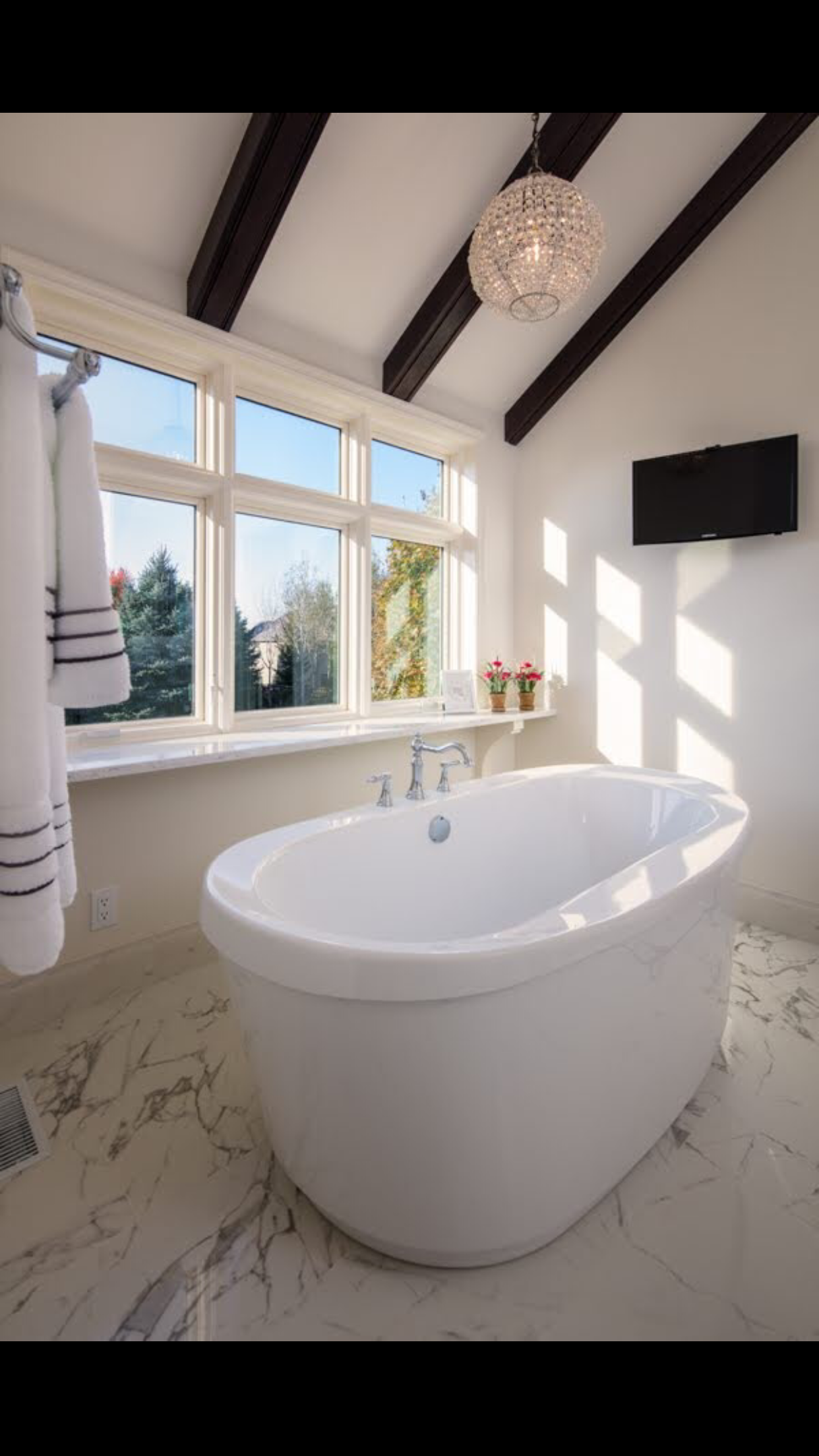 Bathroom with a white freestanding bathtub, large windows with views of trees, hanging chandelier, flat-screen TV on wall, and marble floor.