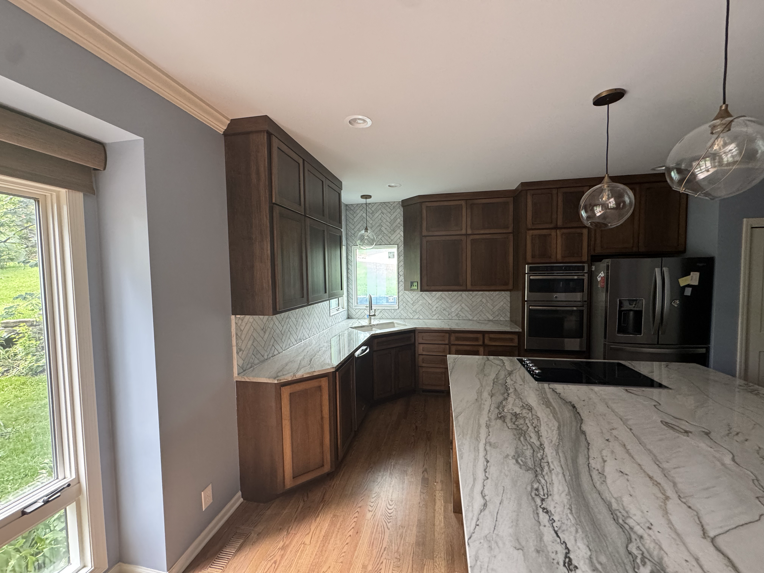 A modern kitchen with dark wooden cabinets, a marble countertop, stainless steel appliances, and pendant lights. There is a large window with greenery outside.