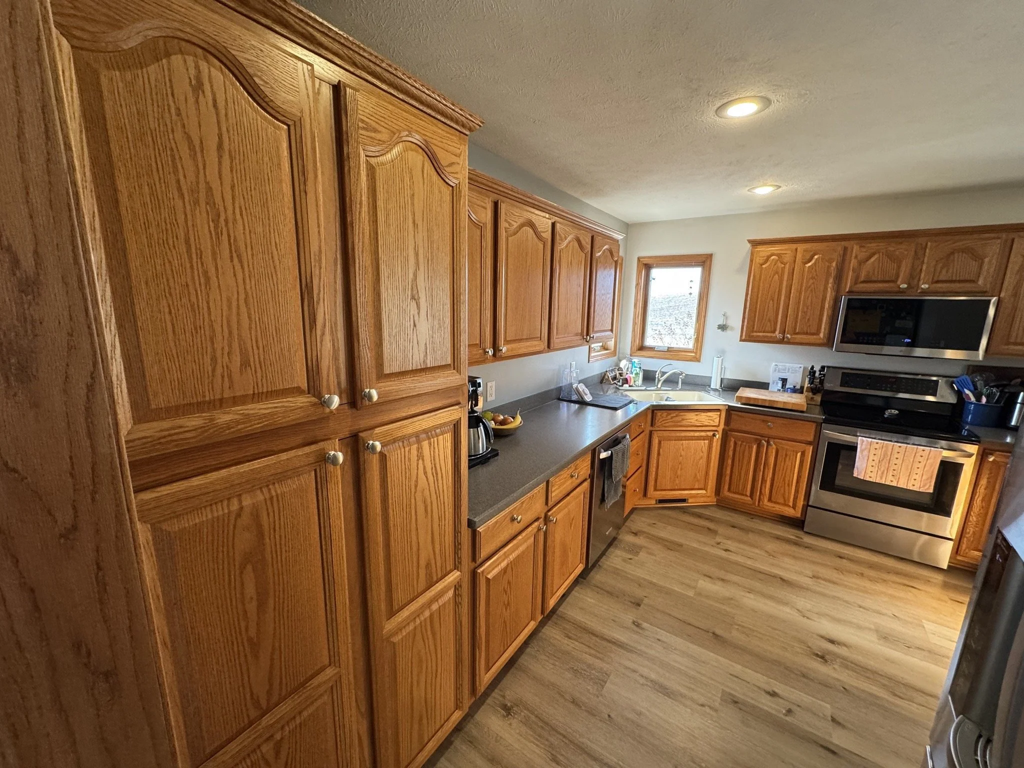 A kitchen with wooden cabinets and a black countertop, featuring a sink, microwave, toaster, and a few kitchen utensils, with hardwood flooring and a small window.
