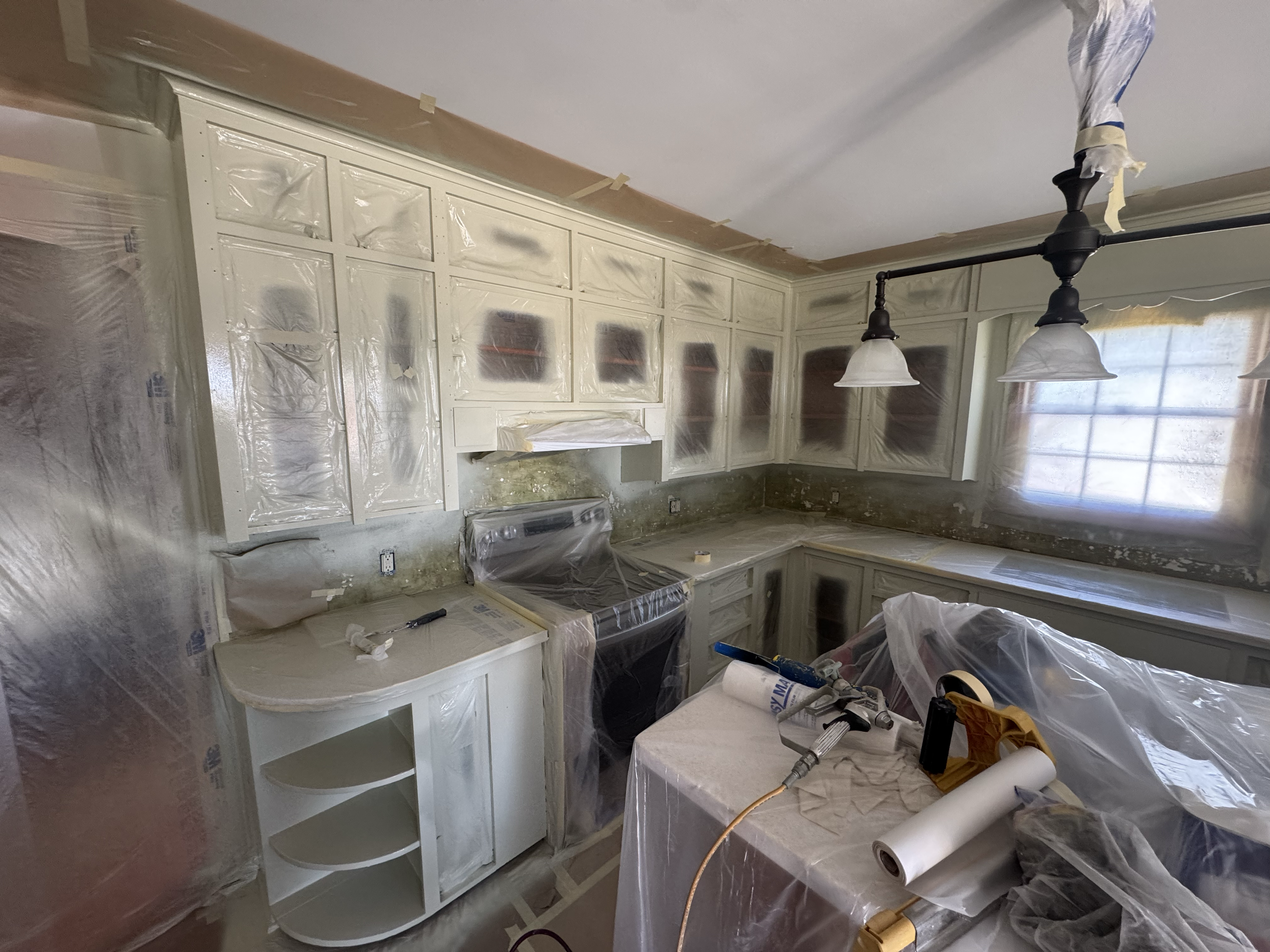 Kitchen cabinets are being painted white and covered with plastic sheeting. The ceiling has tape along the edges for painting. There are tools and materials on the counter, including a roller brush, a spray gun, and a roll of painter's tape. A window