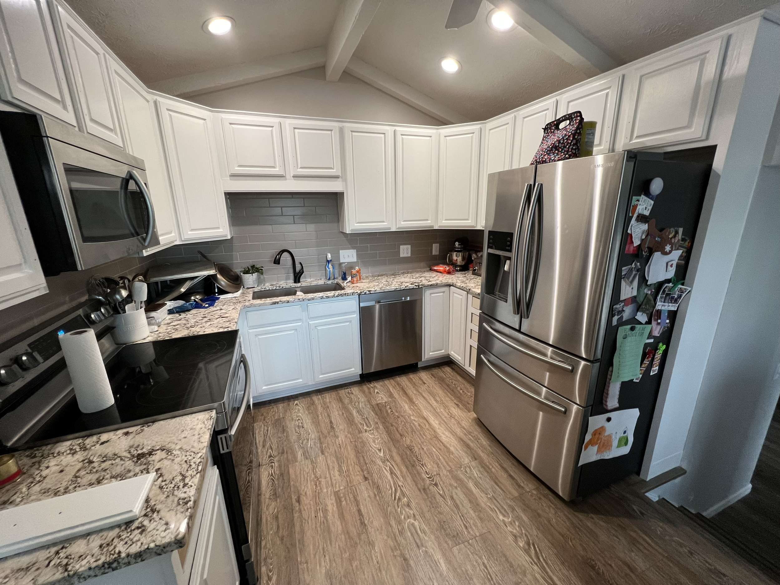 A modern kitchen with white cabinets, granite countertops, stainless steel appliances, and a gray subway tile backsplash. The kitchen features a ceiling with recessed lighting and a wooden floor.