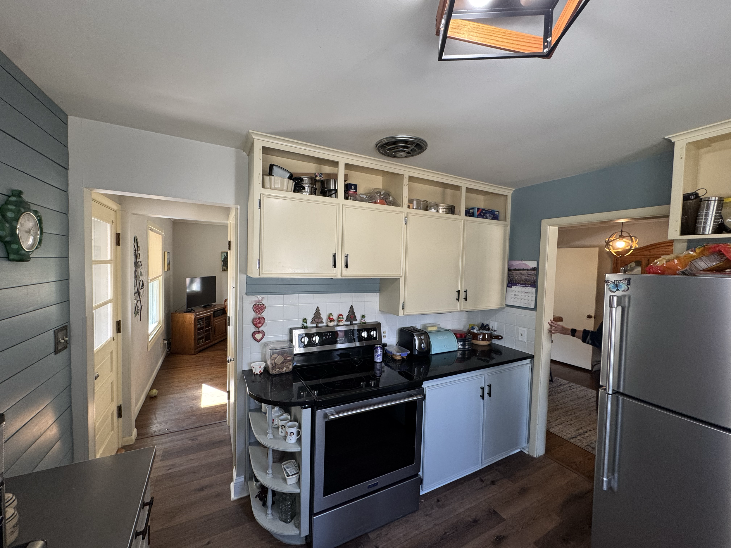 A kitchen with white cabinets, black stove, and wooden shelves. A person is reaching into a refrigerator on the right. The kitchen has wood flooring and a doorway leading to a living room area with a TV and wooden cabinet.