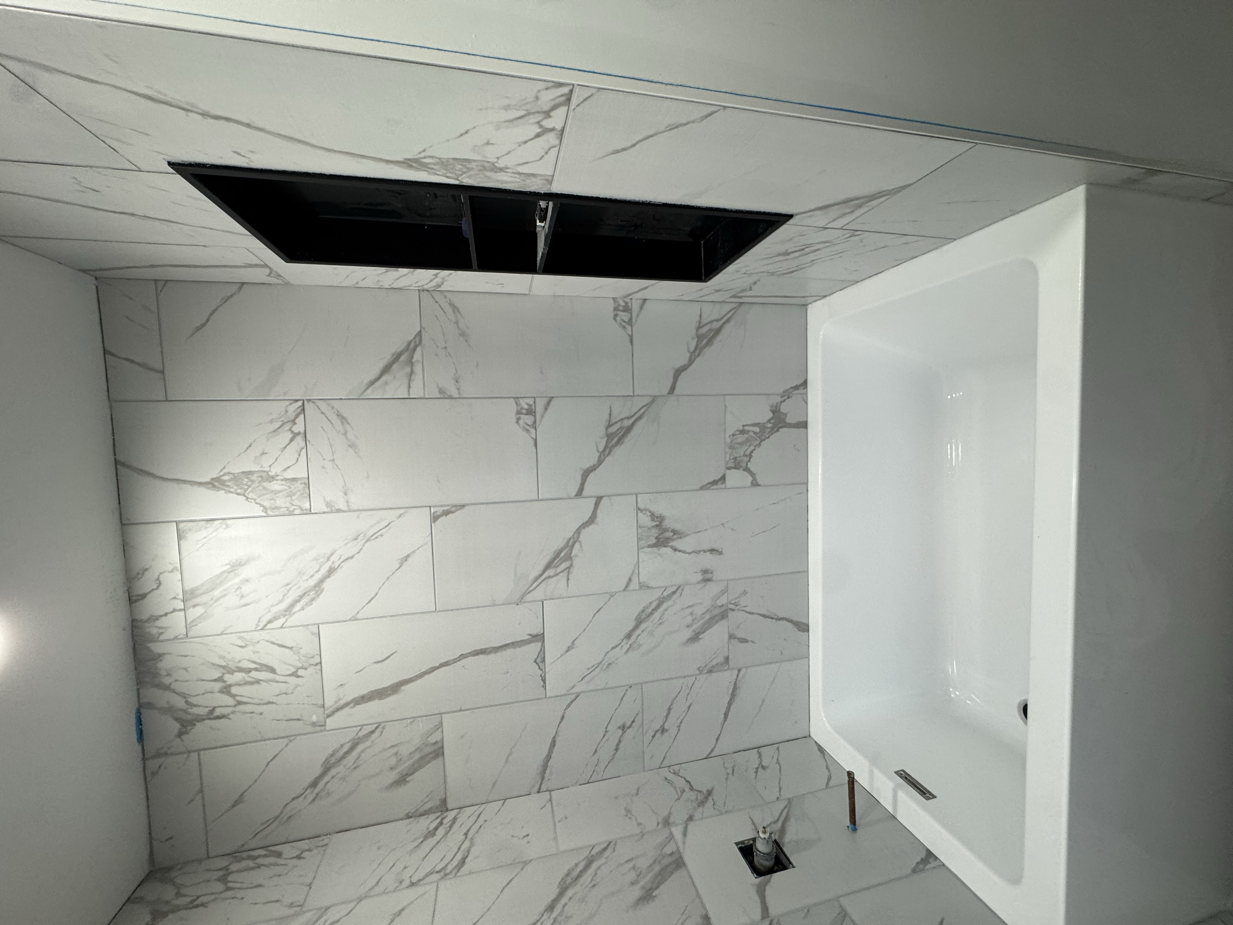 The image shows a bathroom with a marble wall and floor tiles, a wall-mounted sink, and an open cavity in the wall possibly for installing a fixture or storage. There are visible plumbing pipes and a vent or drain opening at the floor.