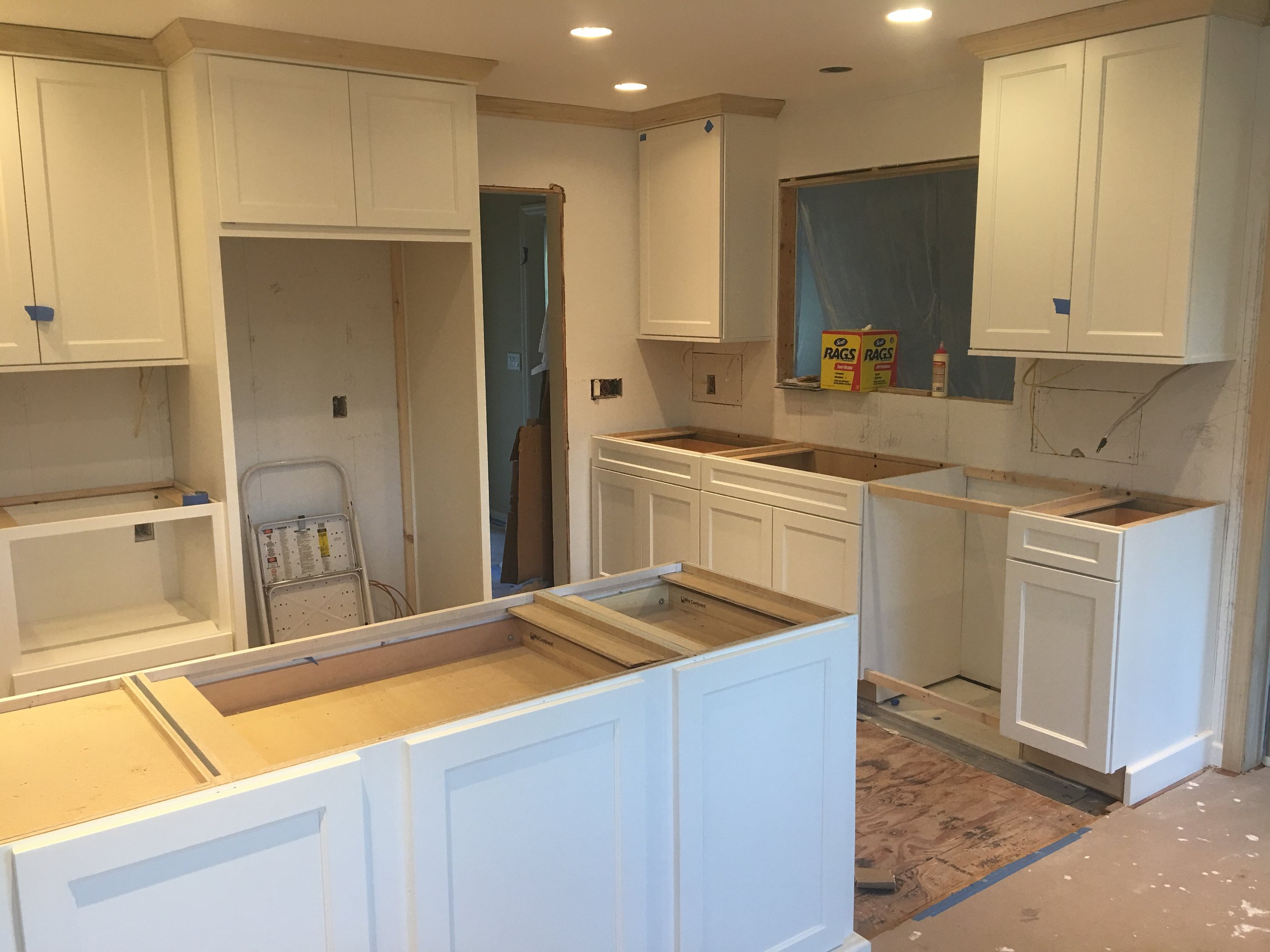 Kitchen cabinets are being installed, with some cabinets missing doors and drawers, and construction materials and tools visible.