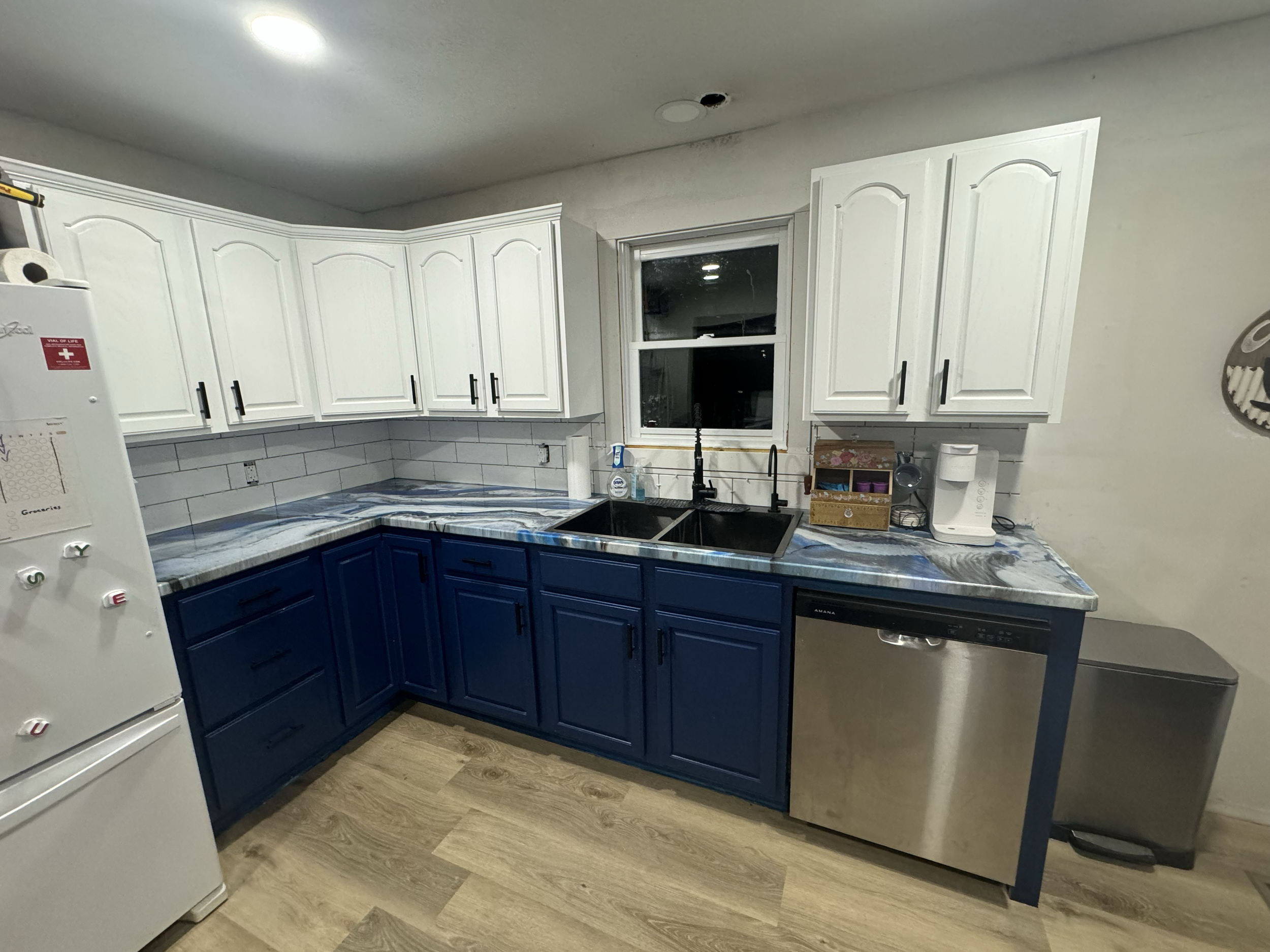 A kitchen with white upper cabinets and blue lower cabinets, a marble countertop, a double sink, and a window above the sink. There is a white refrigerator on the left and a dishwasher on the right. The floor is wood, and there is a roll of paper tow