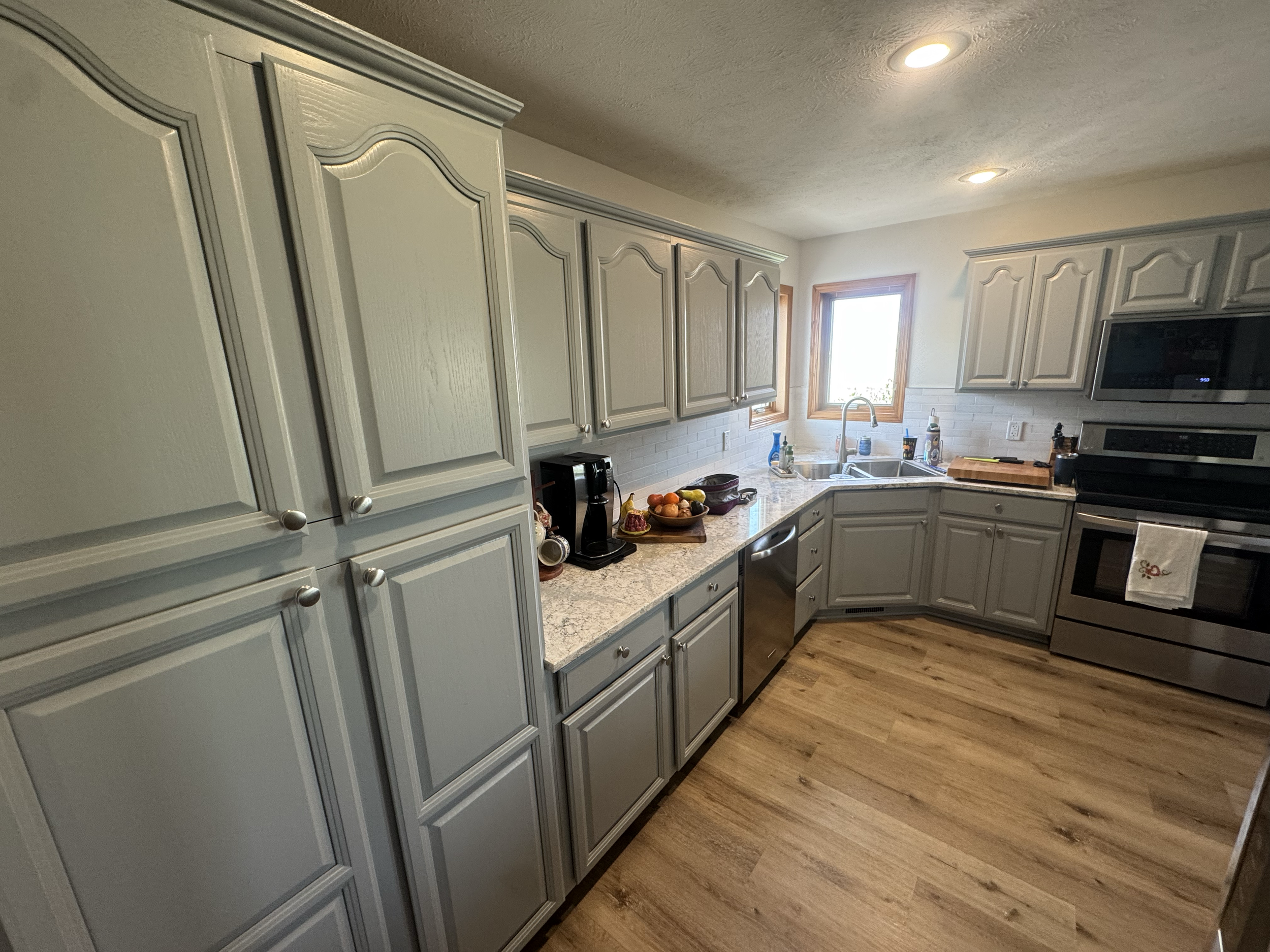 A kitchen with cream-colored cabinet doors, a granite countertop, two windows, and mounted microwave and oven appliances. There are some fruits and kitchen items on the counter.