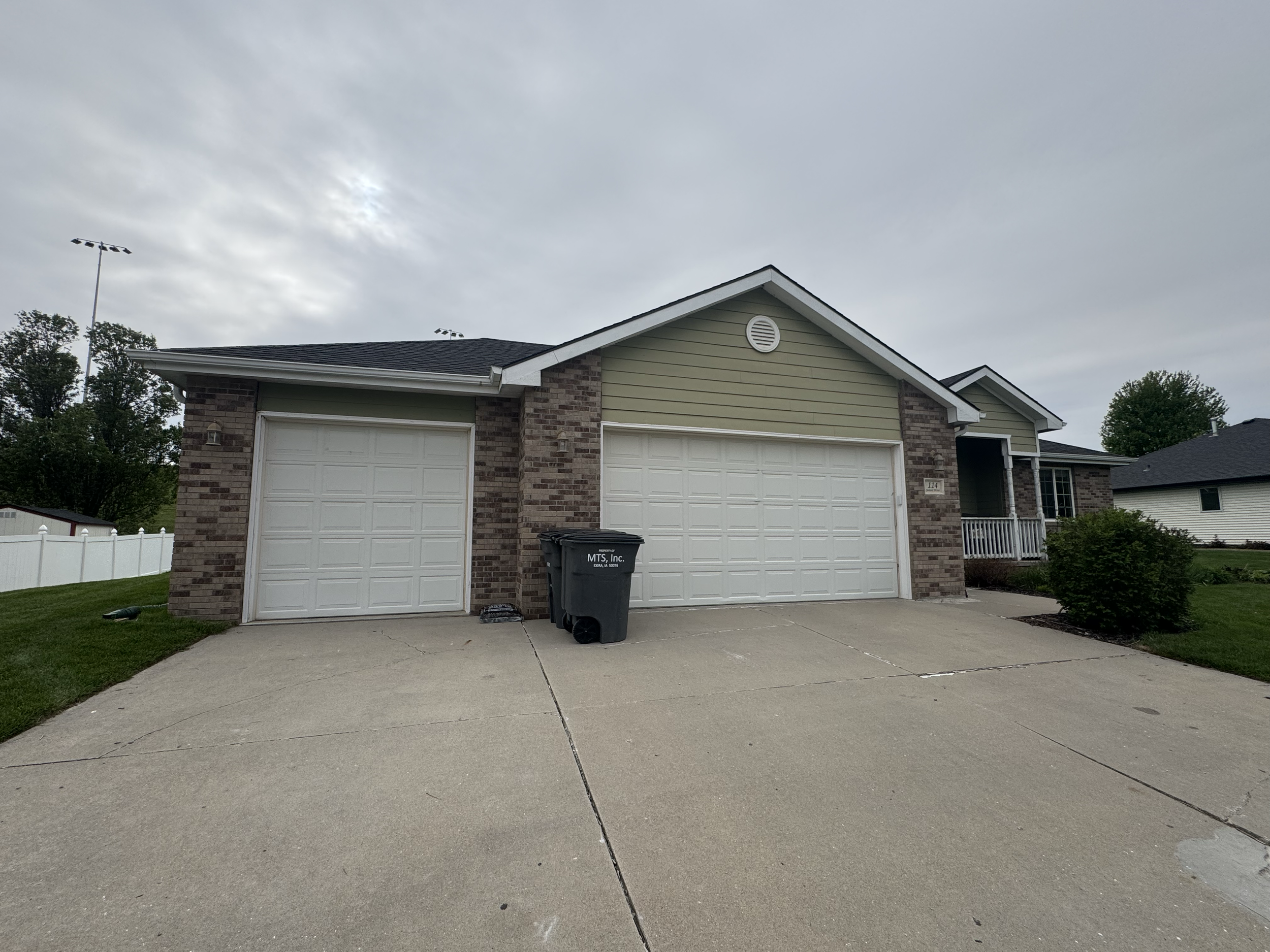 Front view of a suburban house with a two-car garage, green siding, brick accents, a black trash bin in the driveway, green lawn, and trees in the background under an overcast sky.