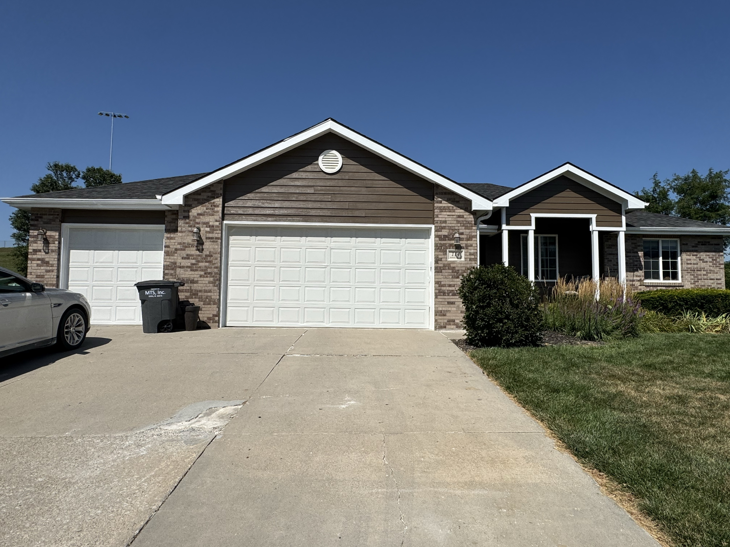 A suburban house with a garage, brick and wooden siding, and a small front porch. There is a concrete driveway with a silver sports car parked on the left, some trash bins in front of the garage, a garden with bushes and grass on the right, and a bri