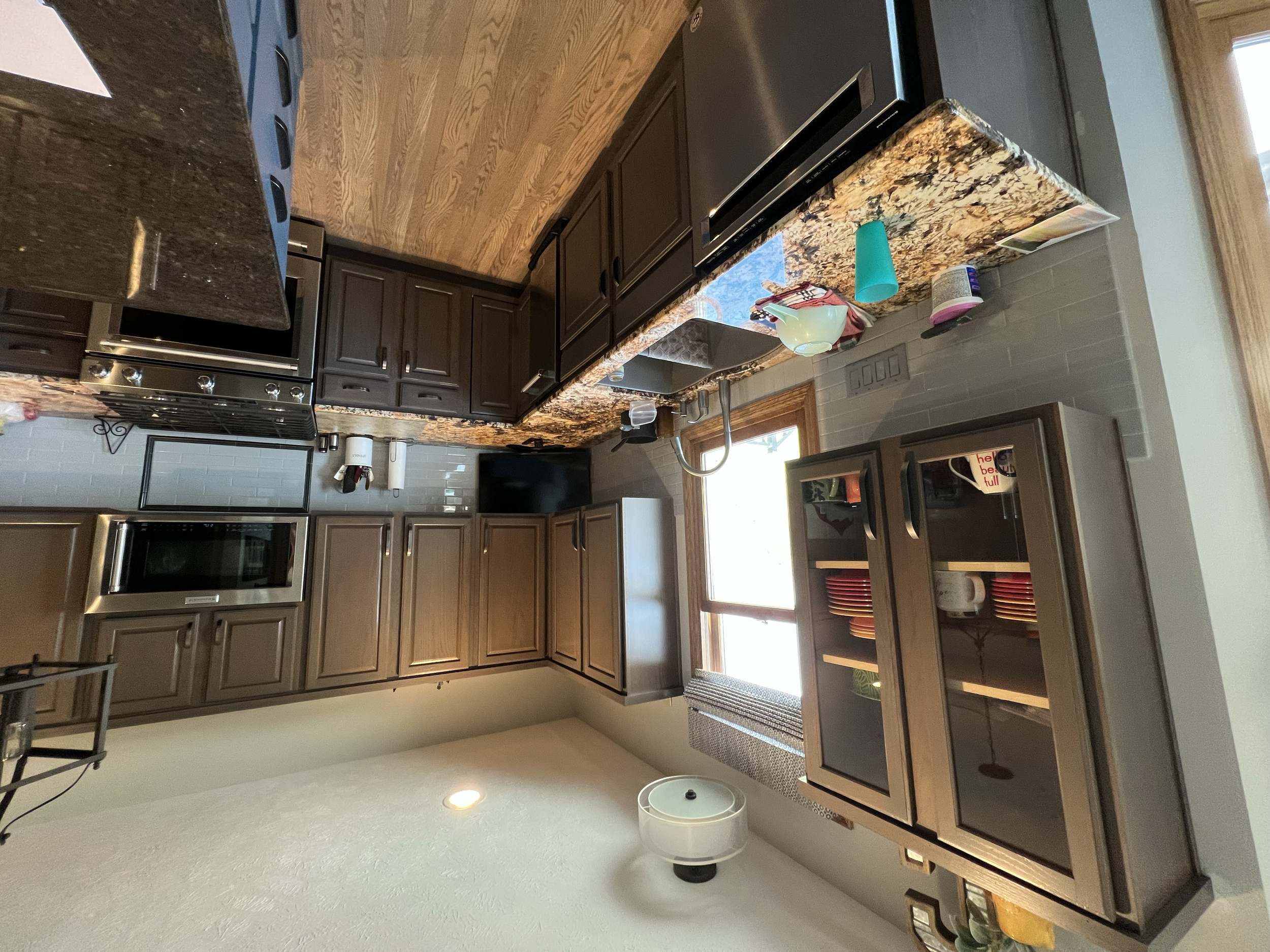 A kitchen with dark brown cabinets, black appliances, granite countertops, a double sink, and a window above the sink. There are various items on the counter including a teapot, paper towel roll, and food containers, with a small trash can on the flo