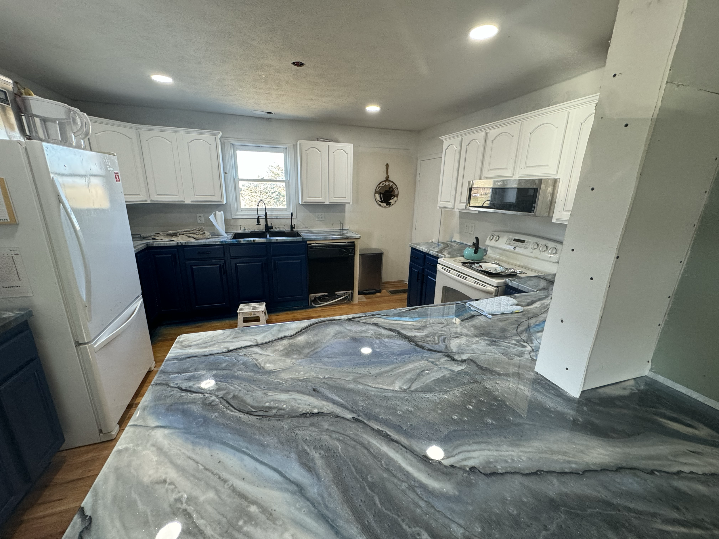 A kitchen under renovation with artificial marble countertops, white upper cabinets, dark lower cabinets, a multi-window over the sink, a microwave above the stove, and various small items on the counters including a turquoise kettle.