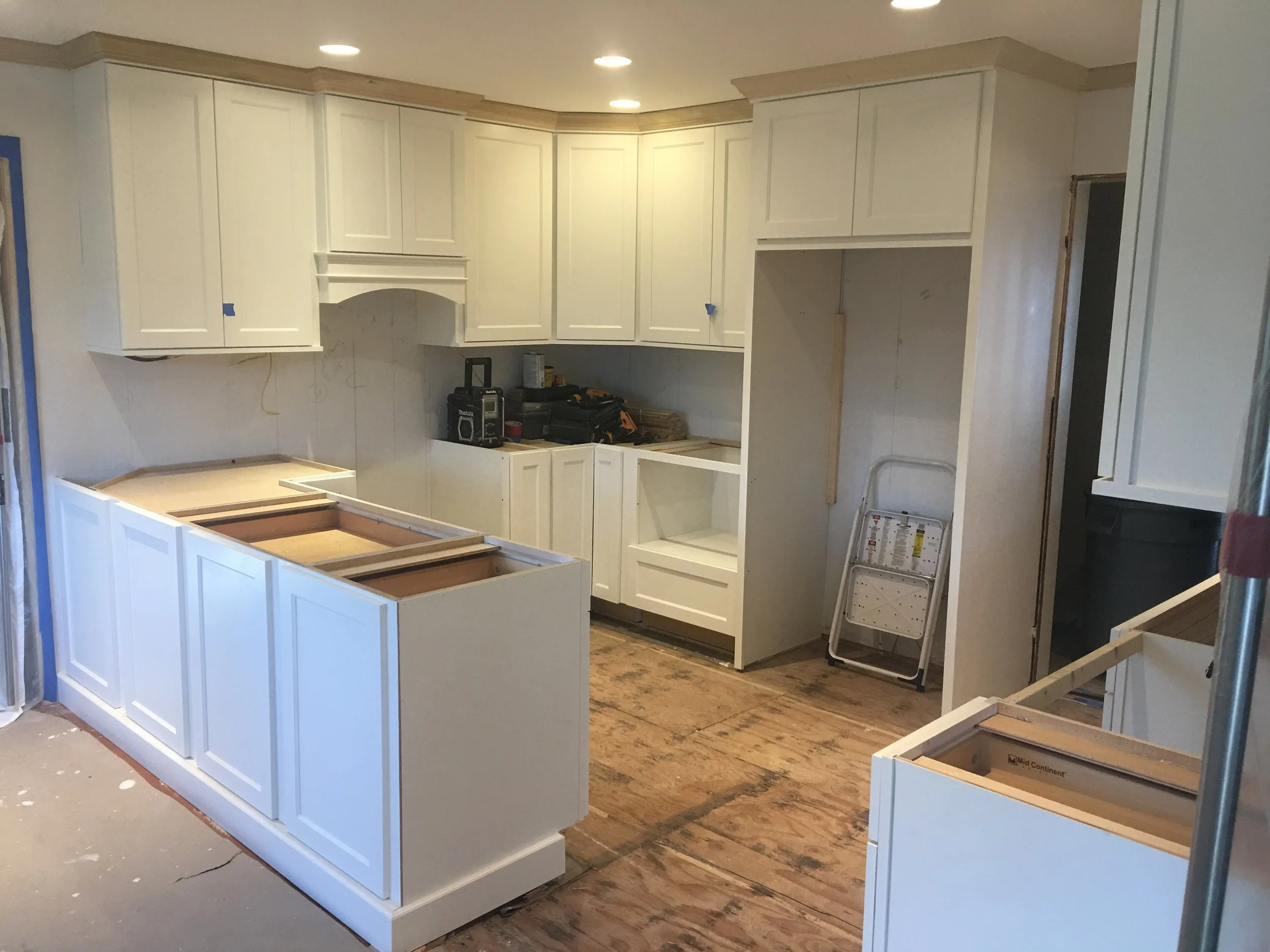 Kitchen cabinets under construction with white painted cabinets and open drawers, construction tools and materials visible, floor unfinished wood, ceiling lights installed.