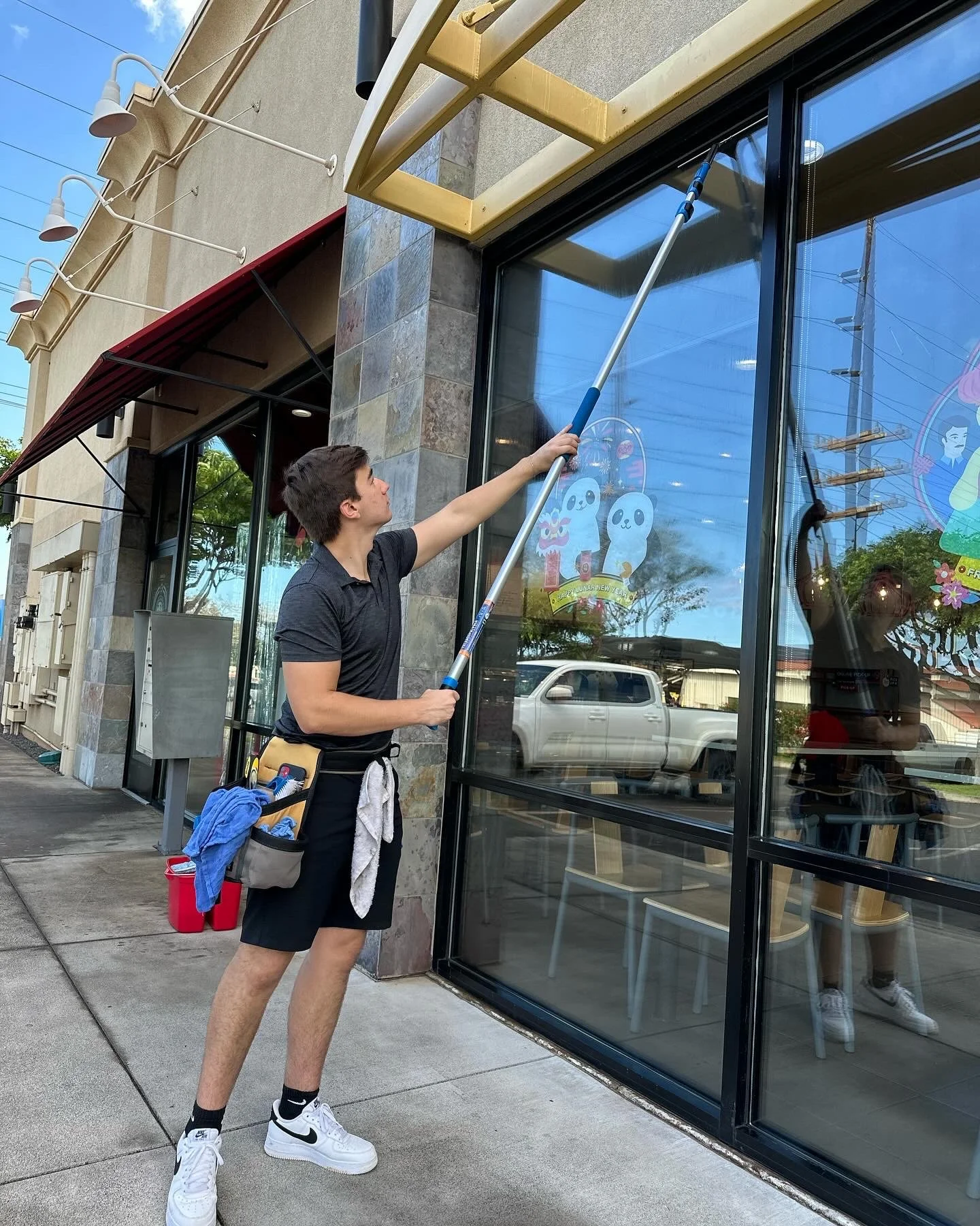 A young man cleaning the large glass window of a building with a long-handled squeegee. The man is wearing a black polo shirt, black shorts, white Nike shoes, and a tool belt with cleaning supplies hanging on his waist. Inside the building, wooden chairs and colorful decals on the window are visible. The sidewalk is wet, and there is a red bucket nearby.