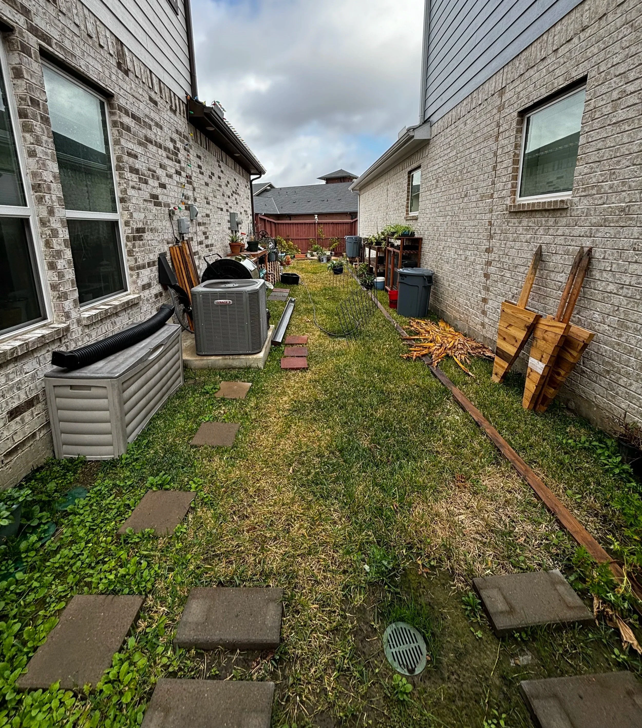 Side yard between two houses with brick exteriors, small grassy area, potted plants, outdoor furniture, and miscellaneous yard items such as wood planks, a lawn mower, and garbage bins.