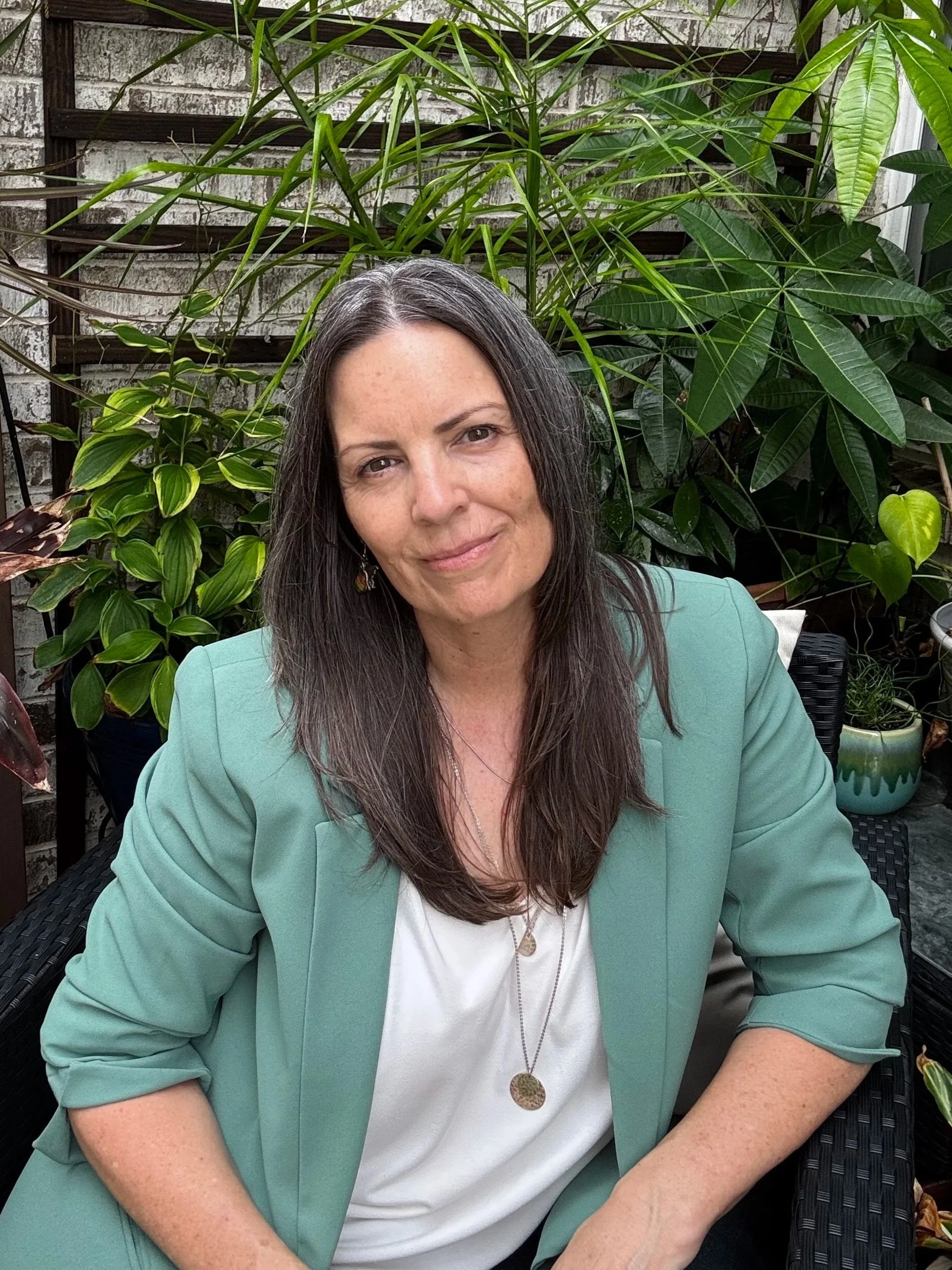A woman with long dark hair, wearing a light green blazer and a white shirt, sitting outdoors with lush green plants and a brick wall in the background.