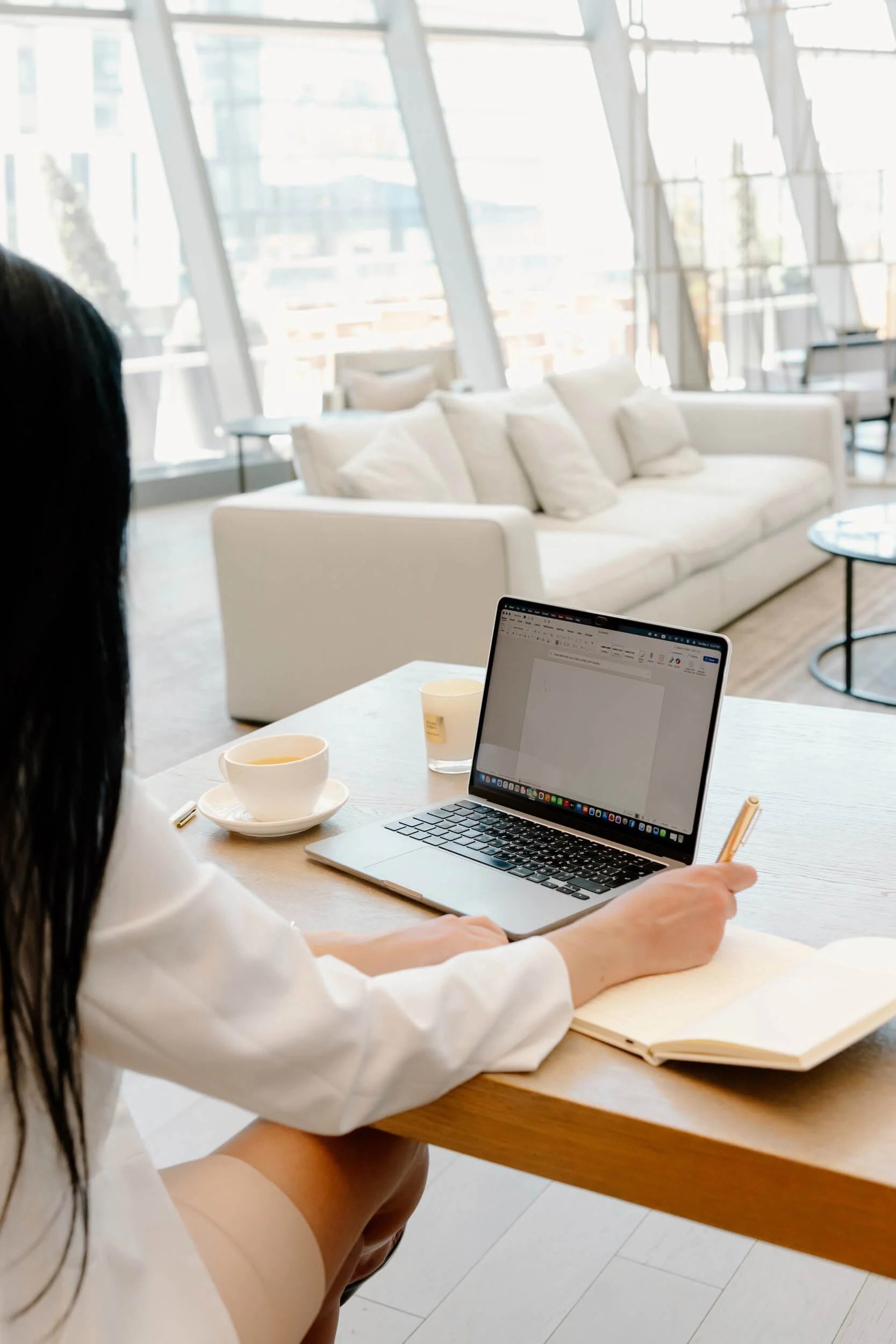 A woman sitting at a wooden table with a laptop, notebook, pens, and coffee cups, working in a bright, modern lounge with large windows and a white sofa in the background.