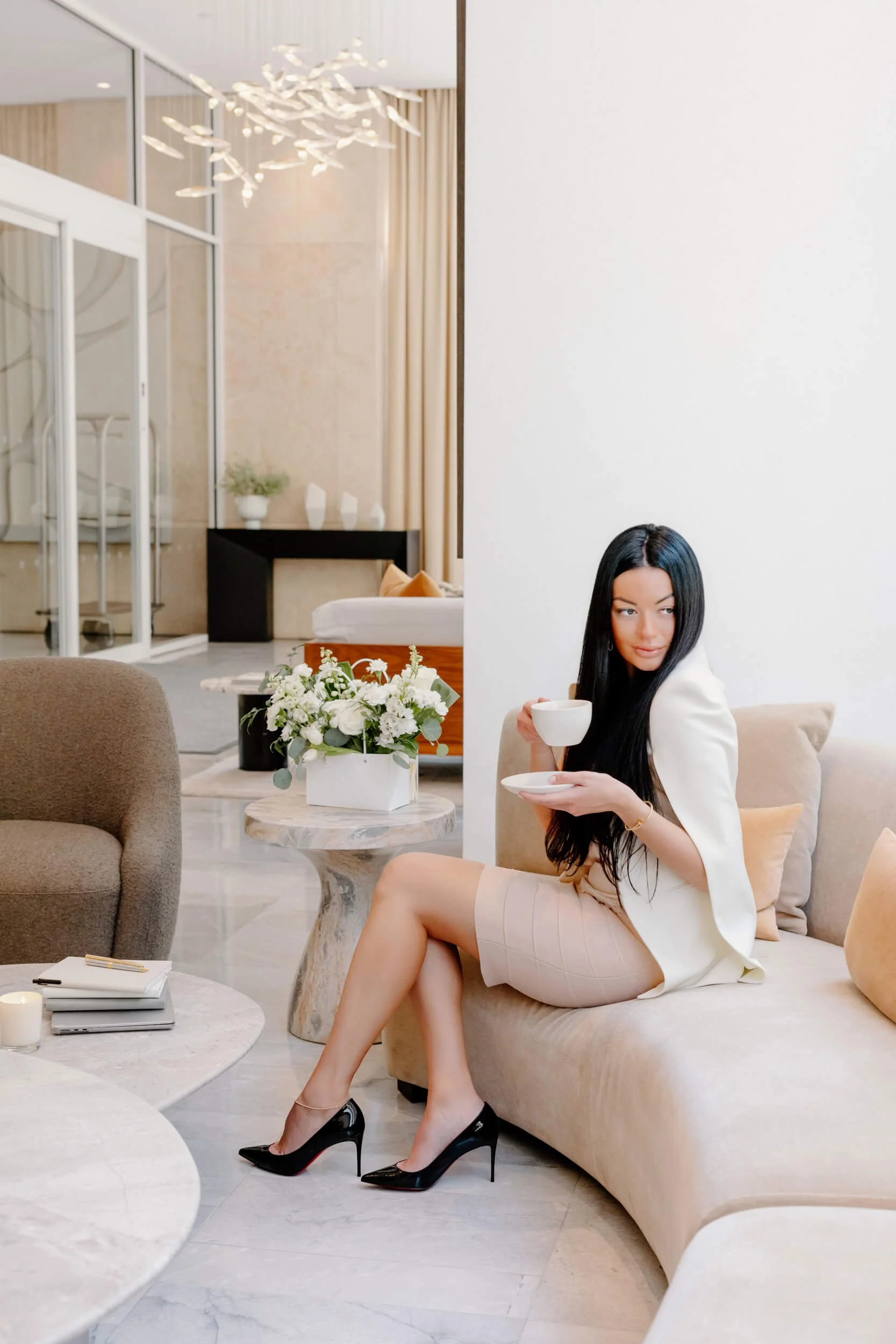 A woman with long black hair, wearing a light pink dress, white blazer, and black high heels, sitting on a beige sofa, holding a teacup and smiling. The room is decorated with white flowers, candles, and modern furniture.