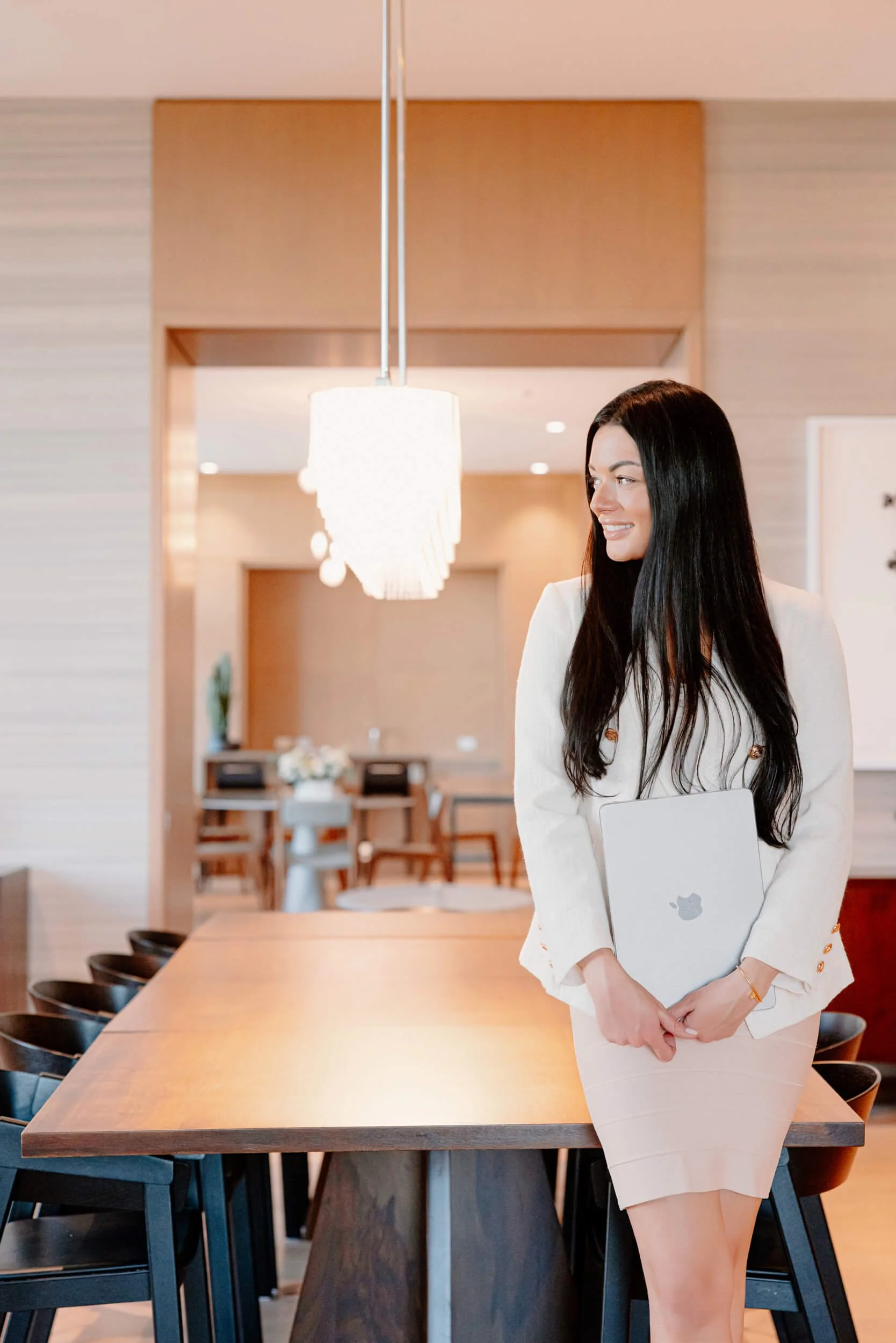 A woman with long dark hair smiling, holding a MacBook laptop, standing in a modern, well-lit conference room with a wooden table and chairs.