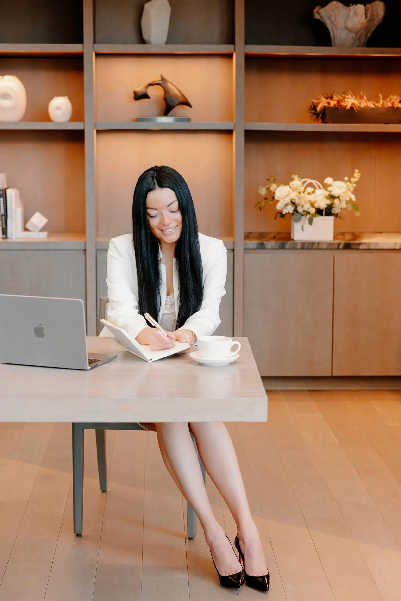 A woman with long black hair getting her nails done, sitting at a desk with a laptop, notebook, and a cup in front of her, in a modern office with wooden decor and decorative objects on shelves.