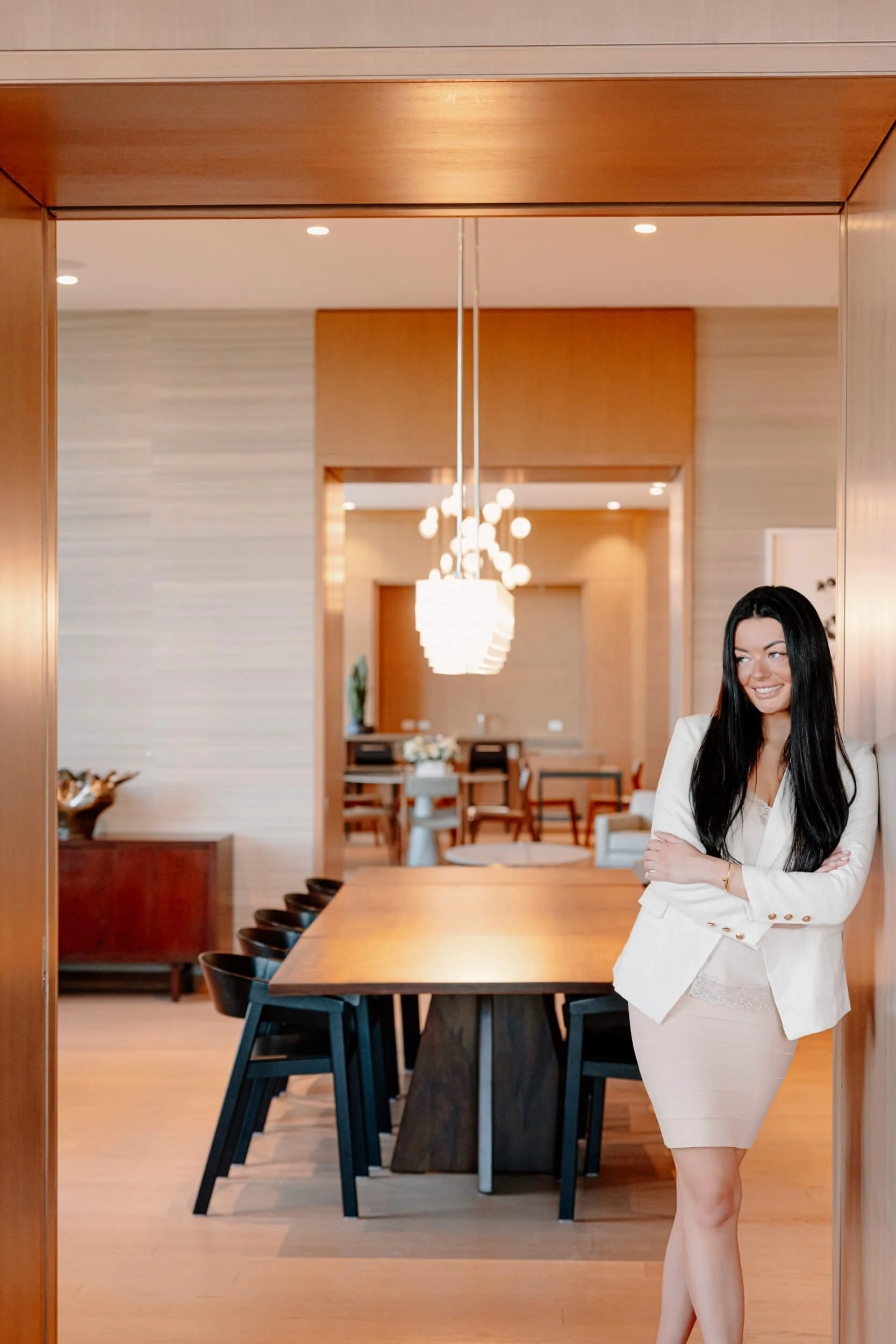 A woman with long black hair wearing a white blazer and skirt, standing with arms crossed in a modern, well-lit dining room with wooden furniture and a large mirror.
