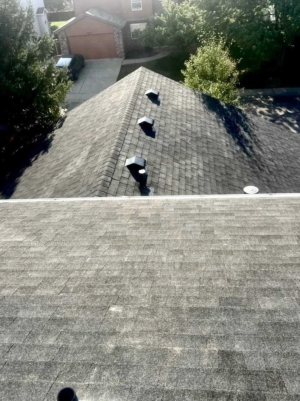 Aerial view of a residential roof with three vent pipes, neighboring rooftops, trees, driveway, and a garage in the background.