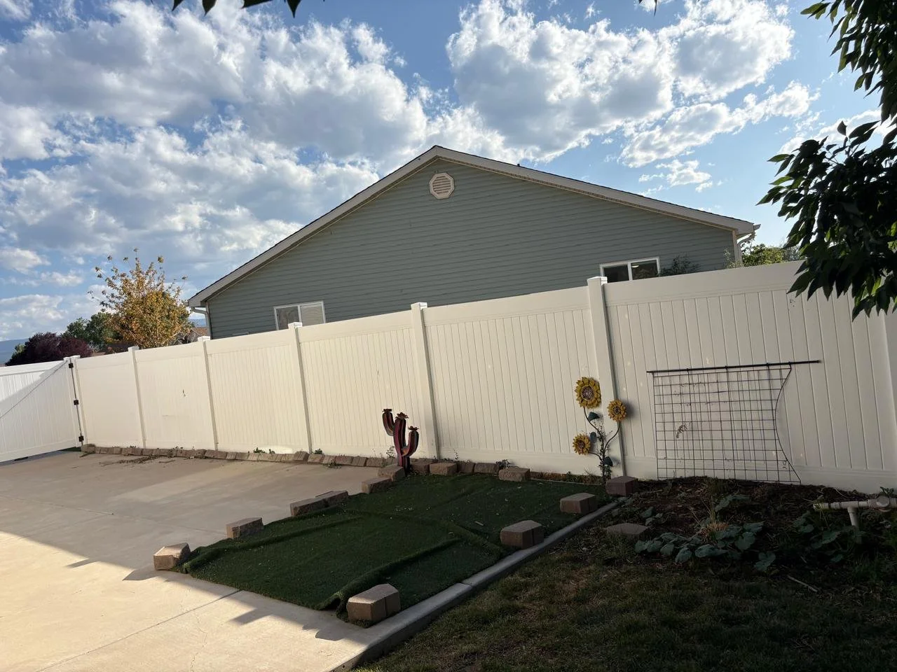 Backyard with a white vinyl fence, house with gray siding in the background, partly cloudy sky, small garden with sunflowers, decorative cactus, small artificial grass patch with bricks, and garden trellis.