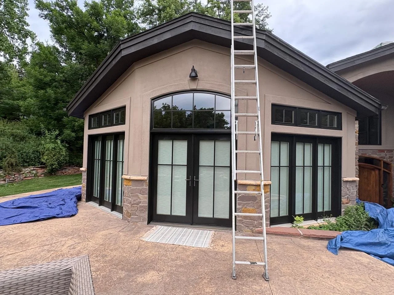 Exterior view of a house with stone accents, black-framed glass sliding doors, and large windows, with a metal ladder leaning against the wall, and some blue tarps on the ground.