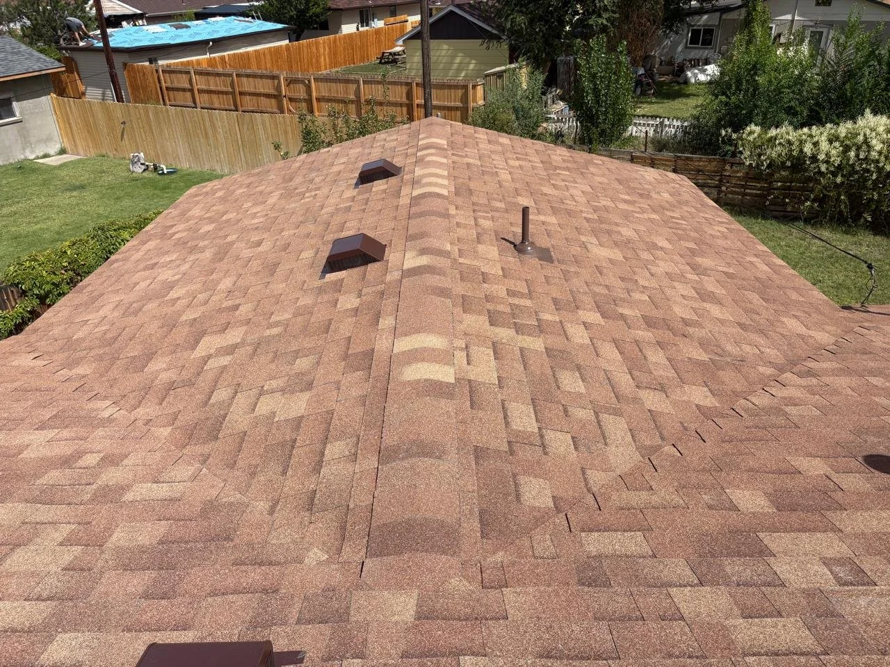 View of a residential house roof with brown asphalt shingles, including two roof vents and a chimney chimney pipe, surrounded by neighboring yards and fences.