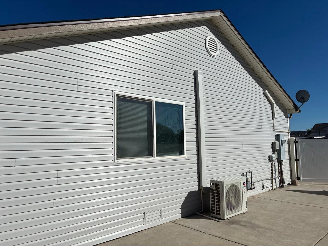 Side of a house with white vinyl siding, a window, an air conditioning unit, pipes, and a satellite dish against a clear blue sky.