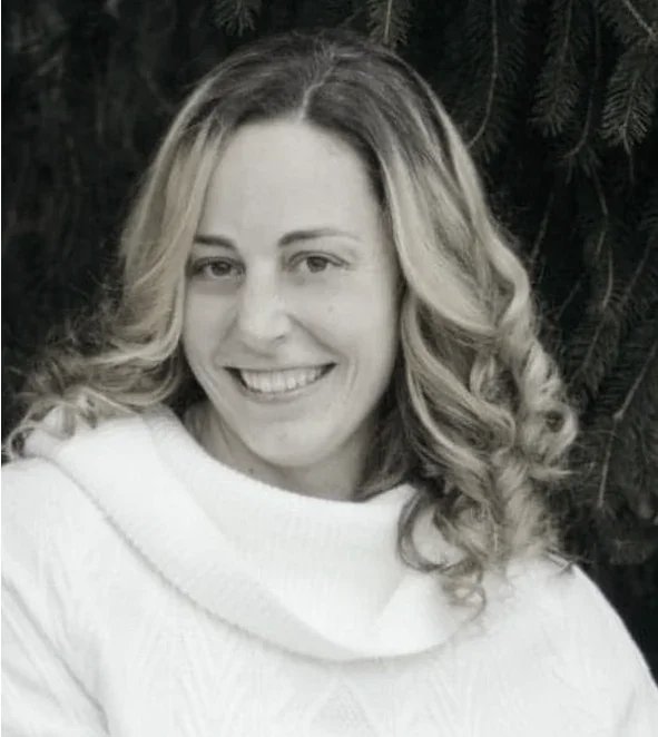 A woman with blonde, shoulder-length, curly hair smiling at the camera, wearing a white sweater with a cowl neck, standing in front of dark tree branches.
