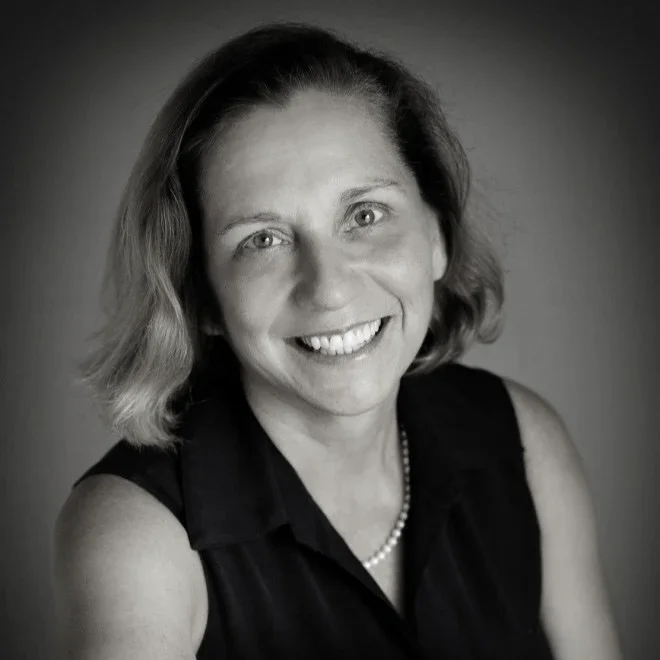 Black and white portrait of a smiling woman with shoulder-length hair, wearing a sleeveless black top and a pearl necklace.