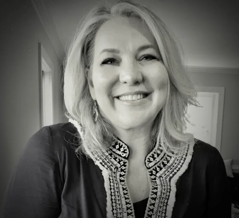 Black and white photo of a woman with shoulder-length blonde hair, smiling at the camera, wearing a dark jacket with embroidered detailing around the collar, indoors.