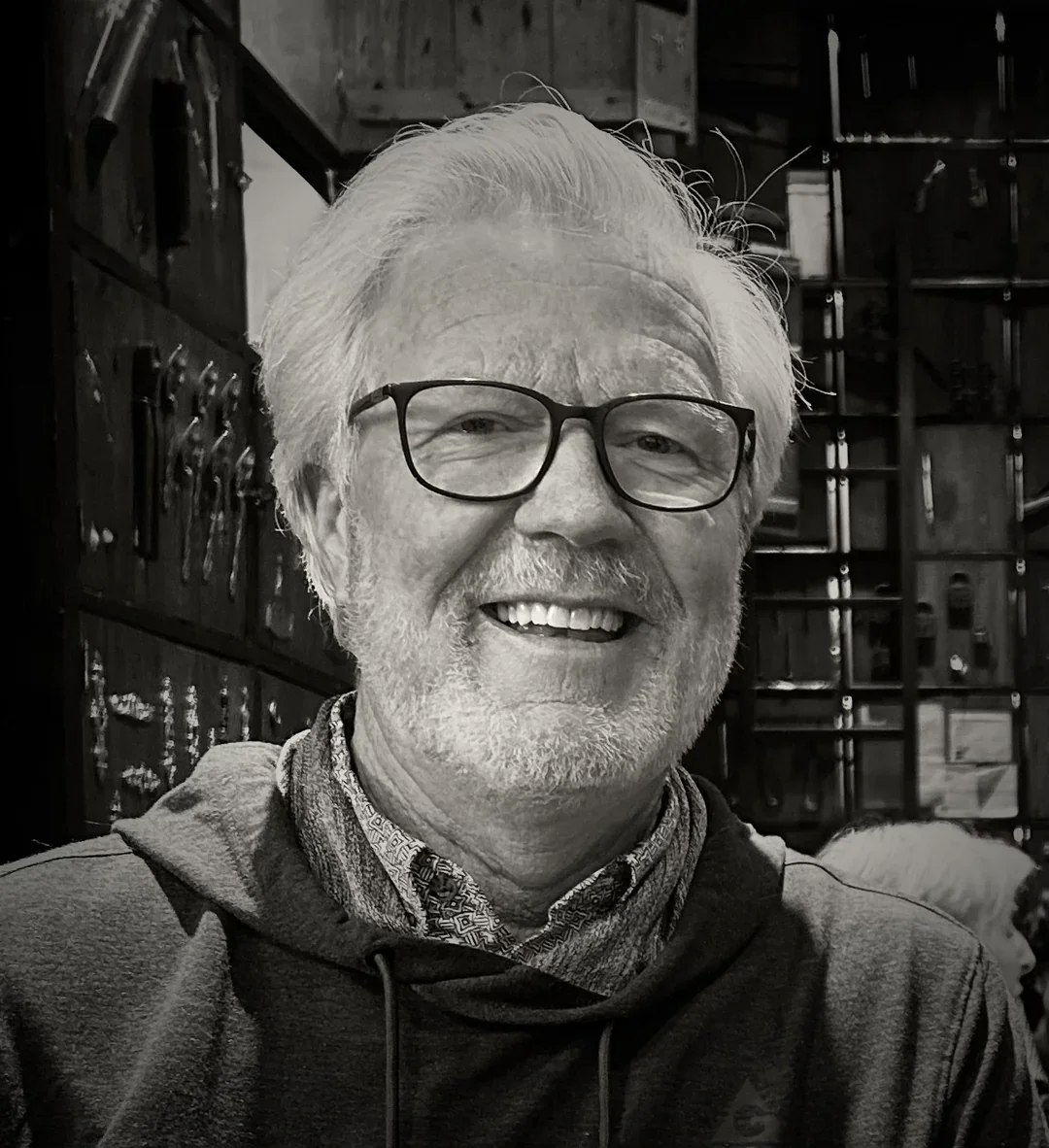 A smiling elderly man with white hair, glasses, and a beard, wearing a hoodie and a patterned scarf, in a workshop or studio with tools on the wall behind him.