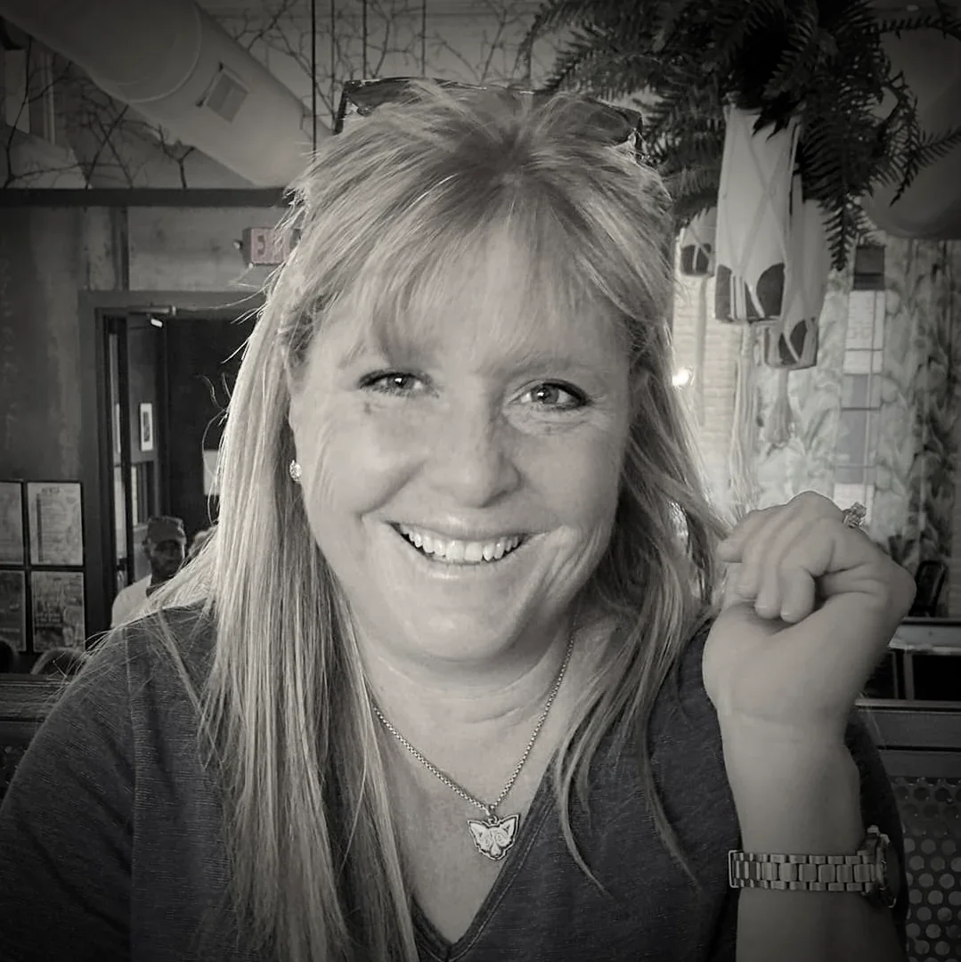A smiling woman with light-colored hair, wearing earrings, a necklace with a butterfly pendant, and a watch, sitting in a restaurant with plants and curtains in the background.