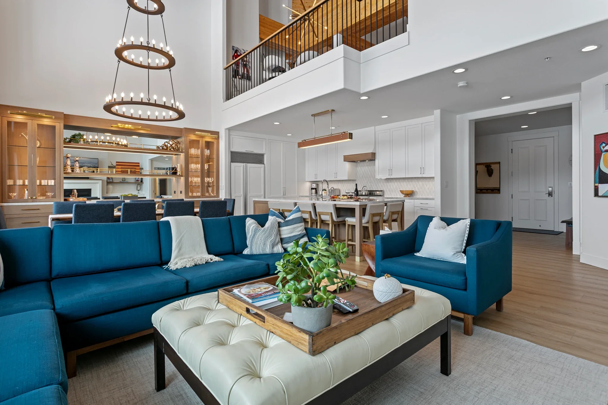 Open-concept living room with blue sofa, decorative pillows, and coffee table with potted plant, overlooking kitchen with white cabinets and island, wooden bar stools, and modern chandelier.