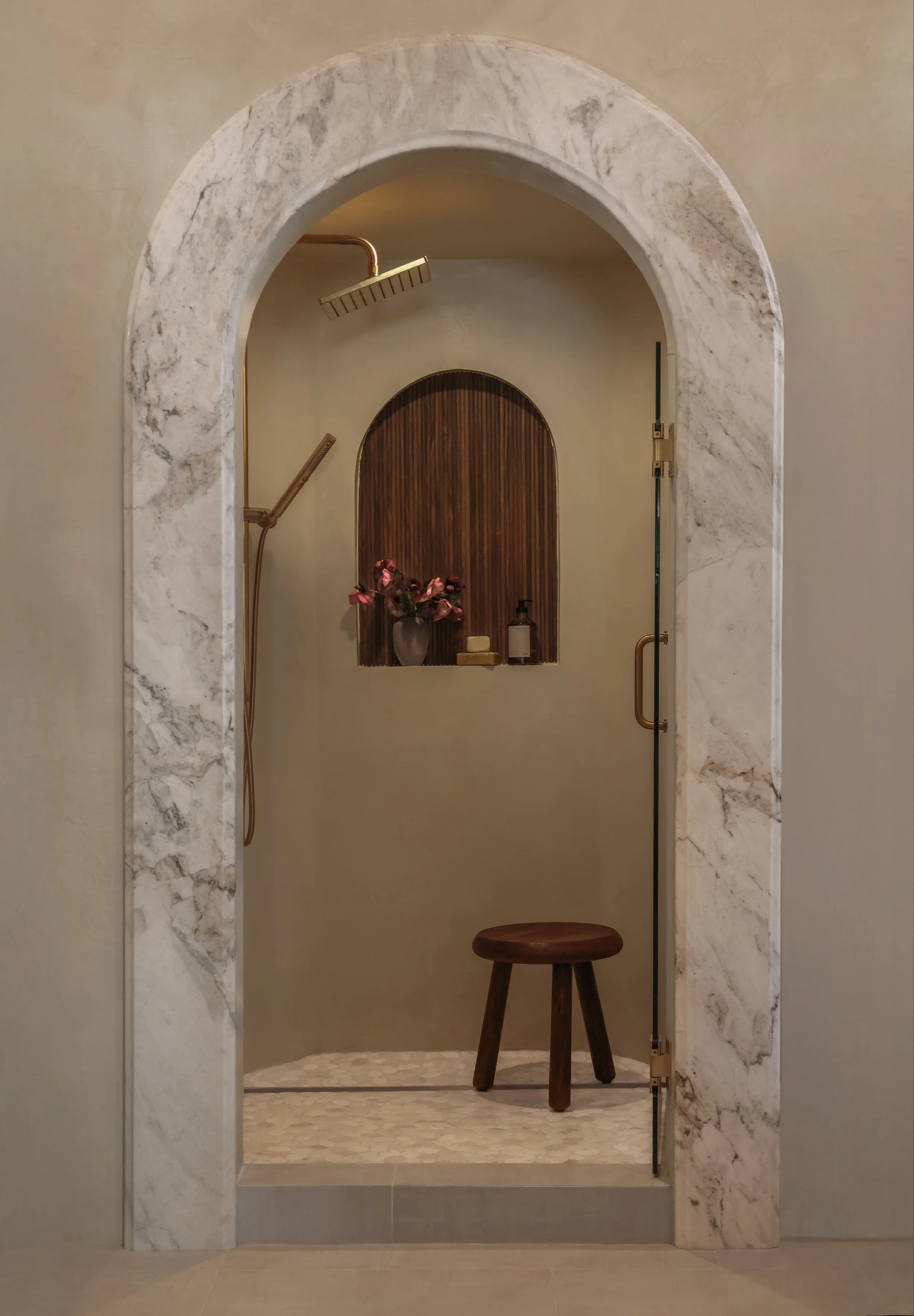 A small wooden stool in front of an arched marble wall leading to a shower area with a gold fixture and a wooden panel. The shower contains a gold showerhead and a gray vase with pink flowers on a shelf, along with a bottle of soap and a soap dish.