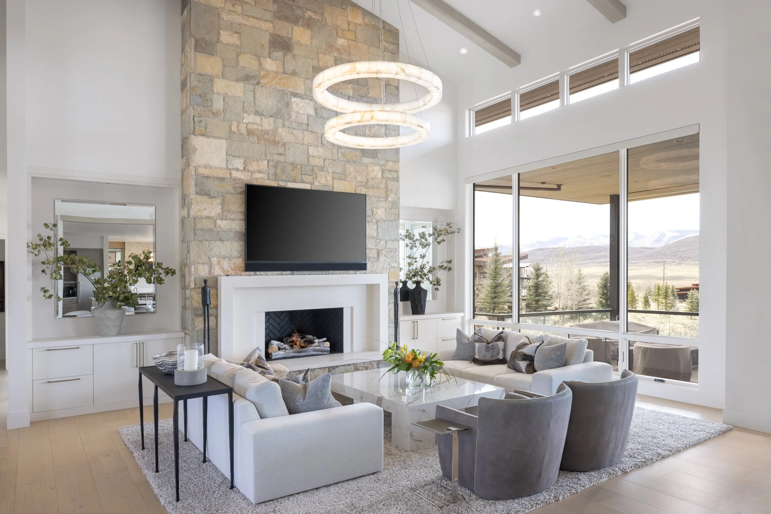 Modern living room with a stone fireplace, a large flat-screen TV, a circular chandelier, white and gray upholstered chairs, and large windows with a scenic view of mountains and trees.