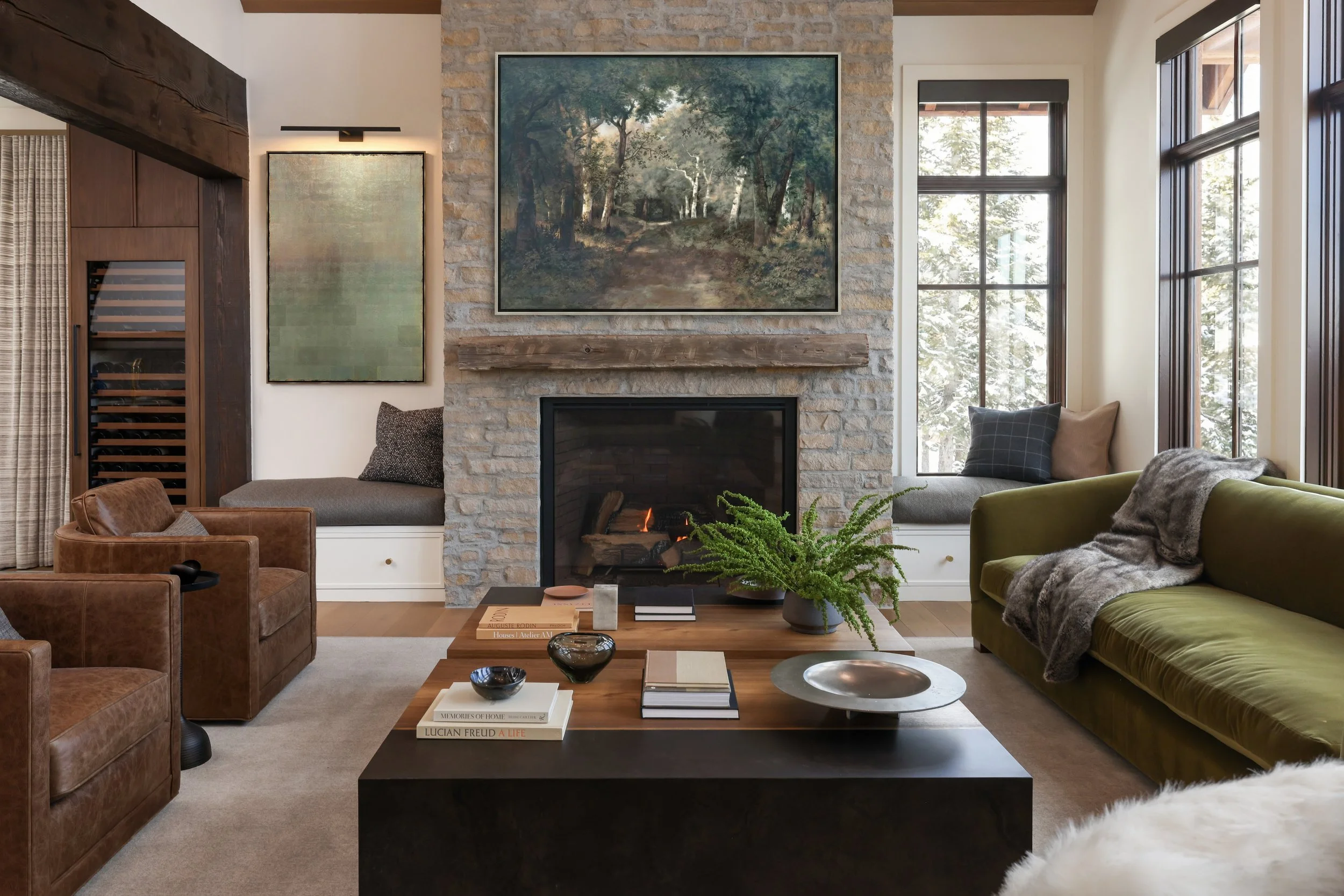 Living room with a stone fireplace, large landscape paintings on the wall, a window seat with cushions, and a coffee table with books and decorative items.