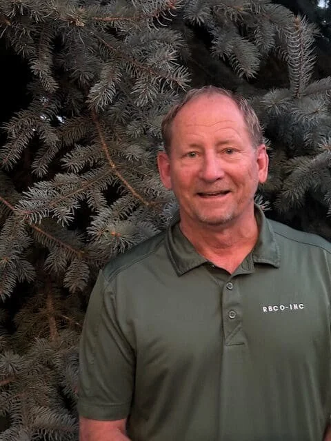 A smiling middle-aged man in a green polo shirt standing in front of pine tree branches.