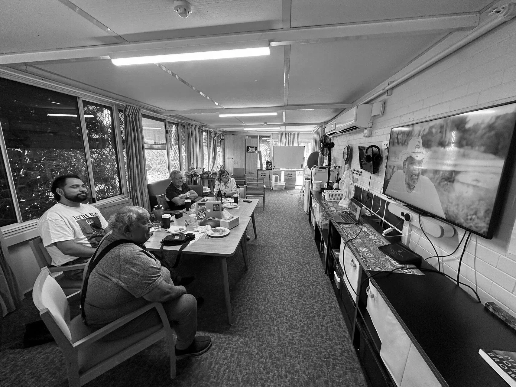 People sitting around a conference table in a room with large windows, some watching a television screen on the wall, with snacks and drinks on the table.
