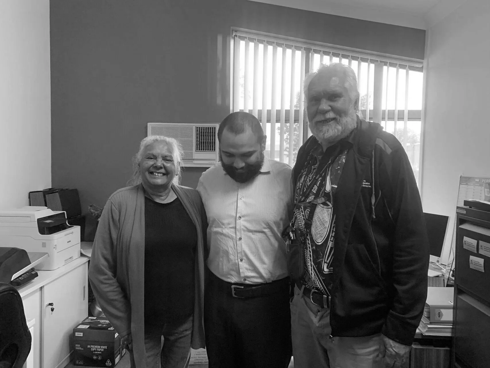 Three people smiling and standing together in an office, with computer equipment and office supplies around them.