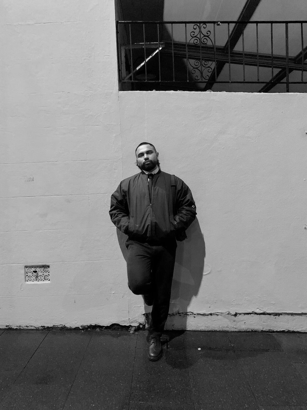 Birrugan Dunn-Velaco standing against a wall, with one foot resting on the wall and hands in his pockets, dressed in a dark jacket and pants. The photo is in black and white.