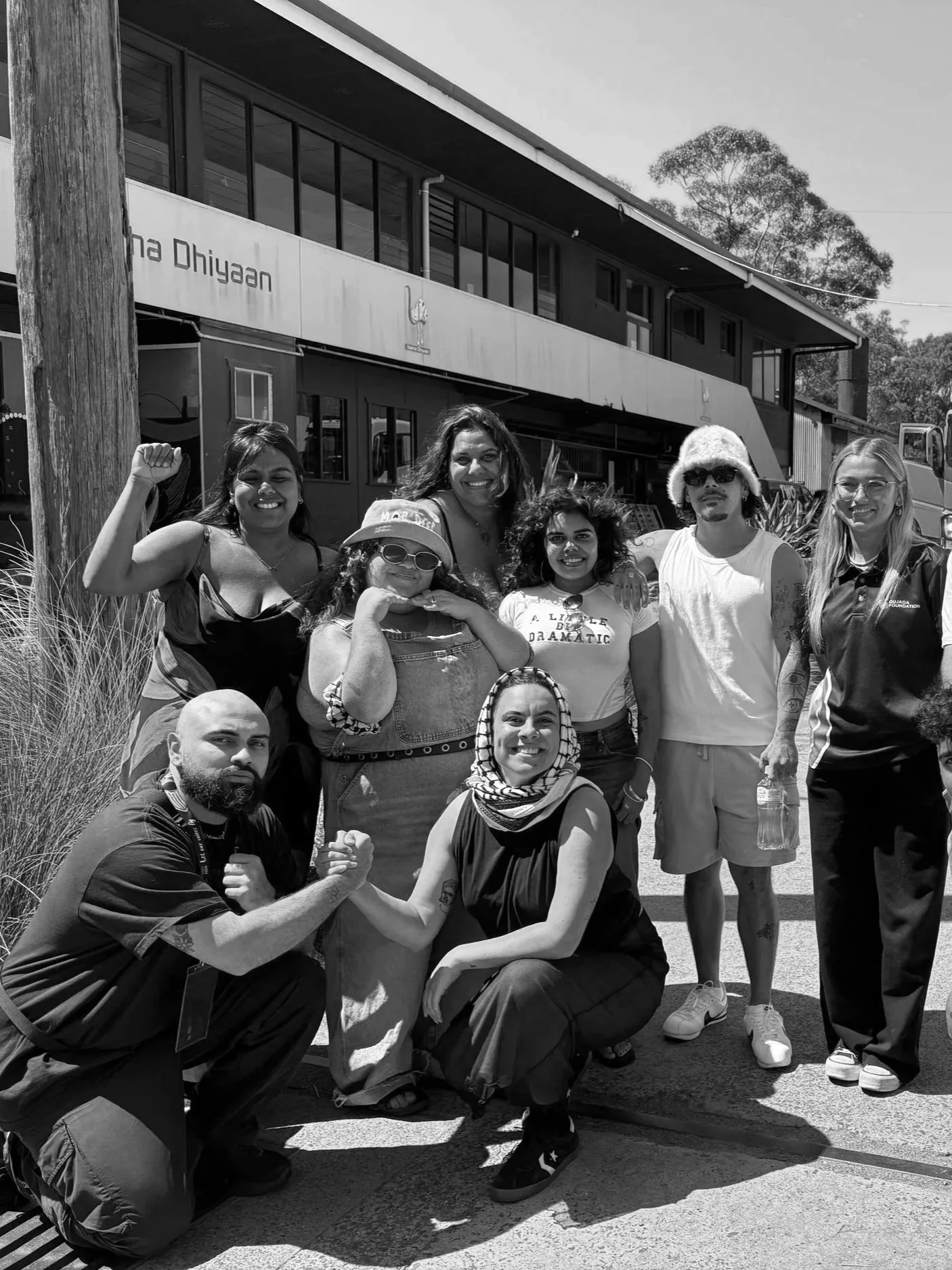 A group of diverse people posing outdoors in front of a building, some showing fists and smiles, with trees in the background.