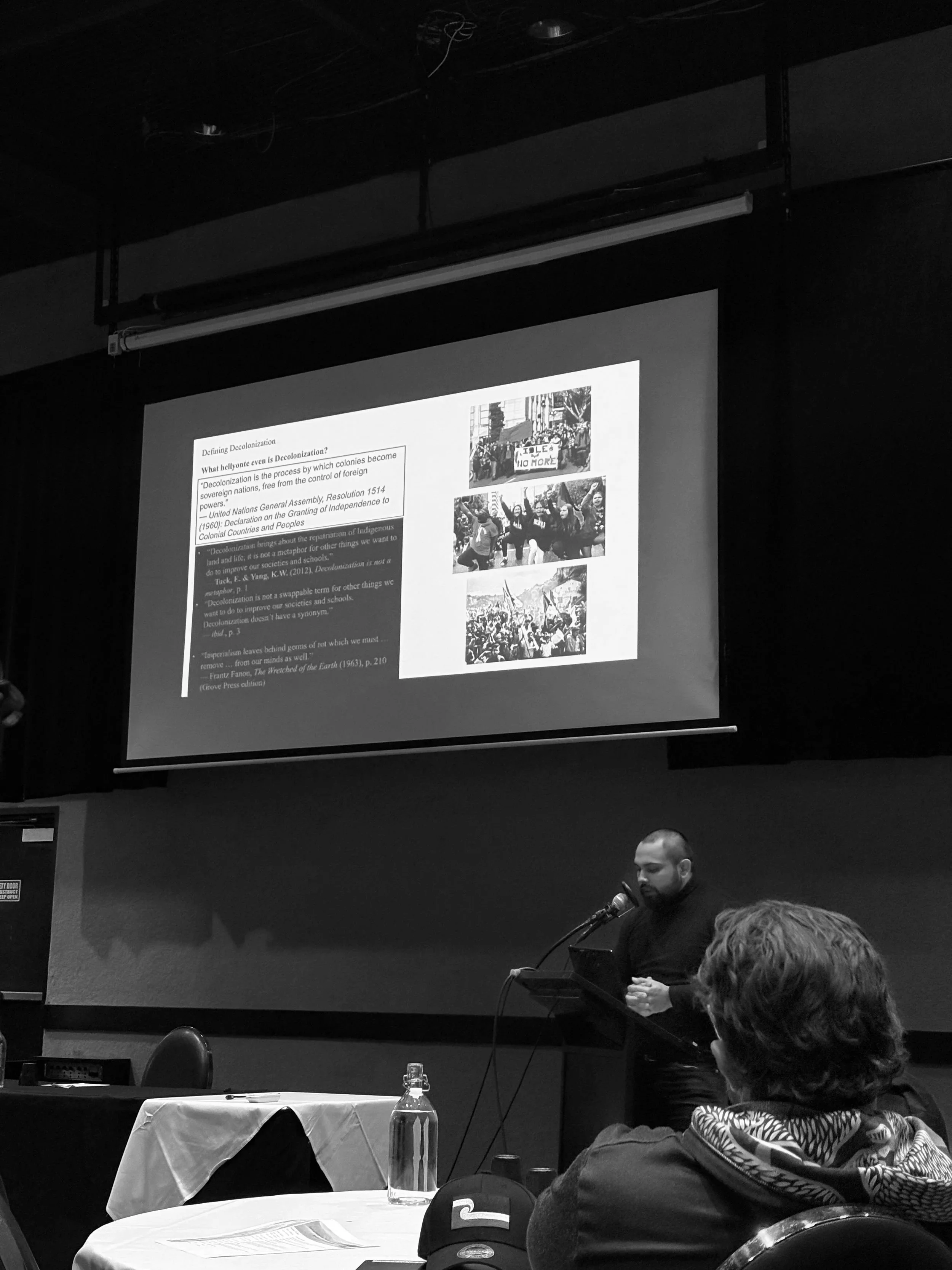 A person is speaking into a microphone at a podium during a presentation or conference. In the background, a large screen displays a slide with text and images related to decolonization. Audience members are visible, seated at tables covered with tablecloths.