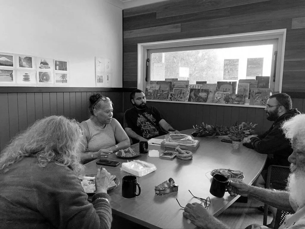 Six people sitting around a wooden cafe table, some eating and drinking, with books and plants on the windowsill, in a cozy room with wood-paneled wall.