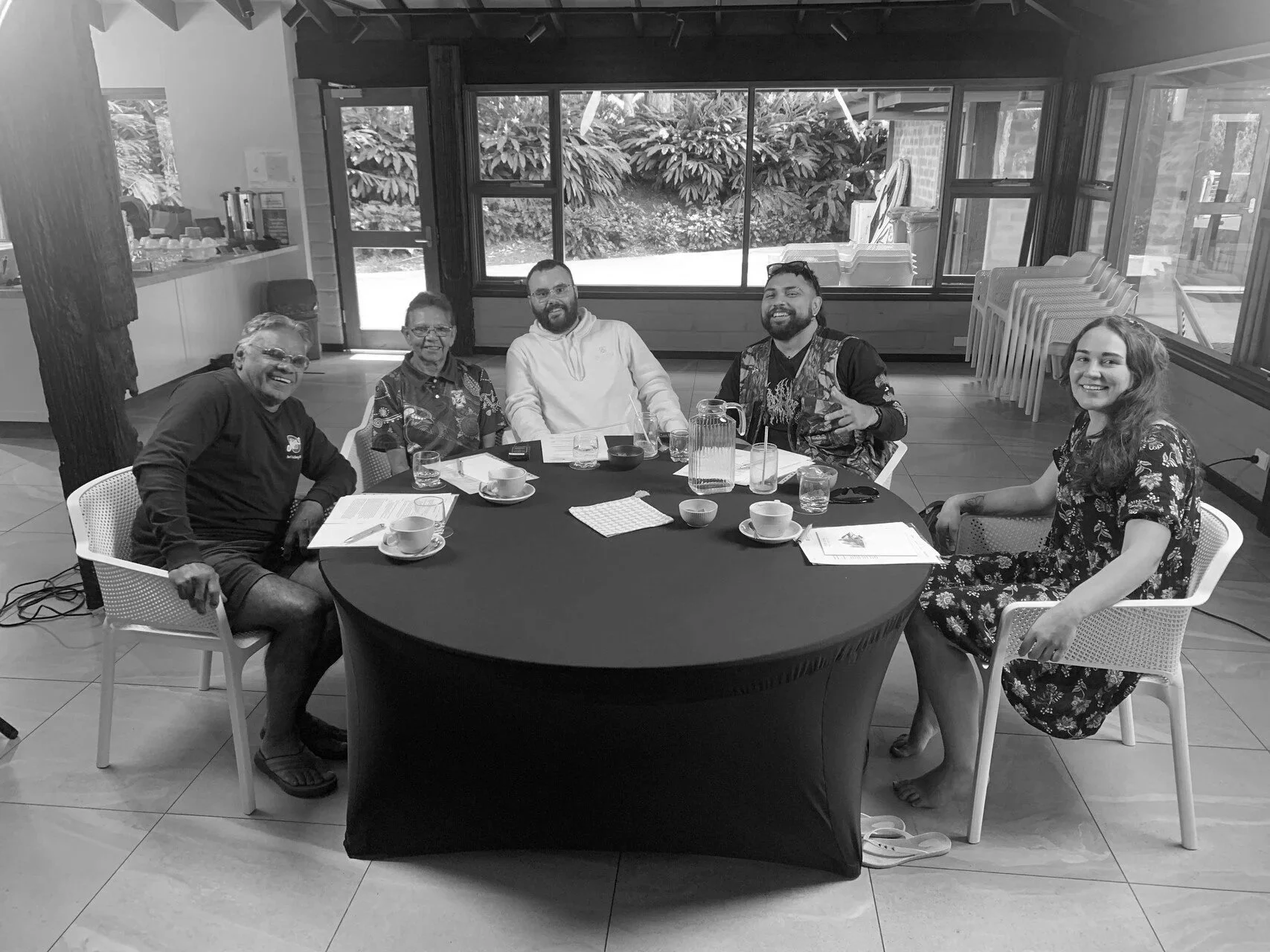 Six people sitting around a table in a bright, indoor setting, smiling at the camera. The table has cups, glasses, and papers. Large windows show foliage outside.